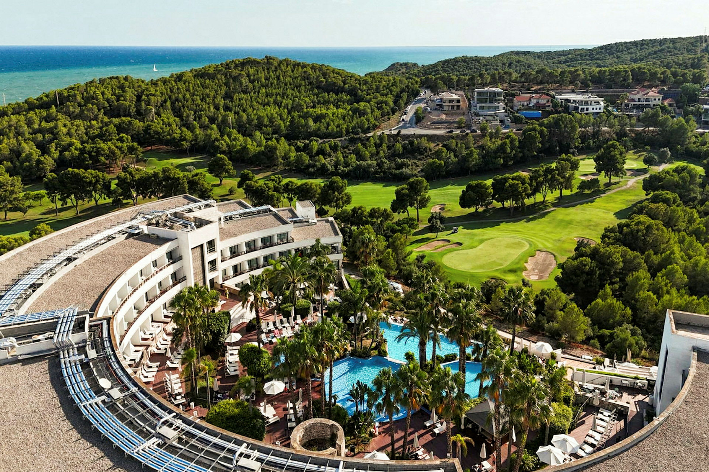 Bild av en hotellbyggnad med pool och omgivande palmträd, omgivet av gräsmattor och skog med havet i bakgrunden. (AI-genererad text)