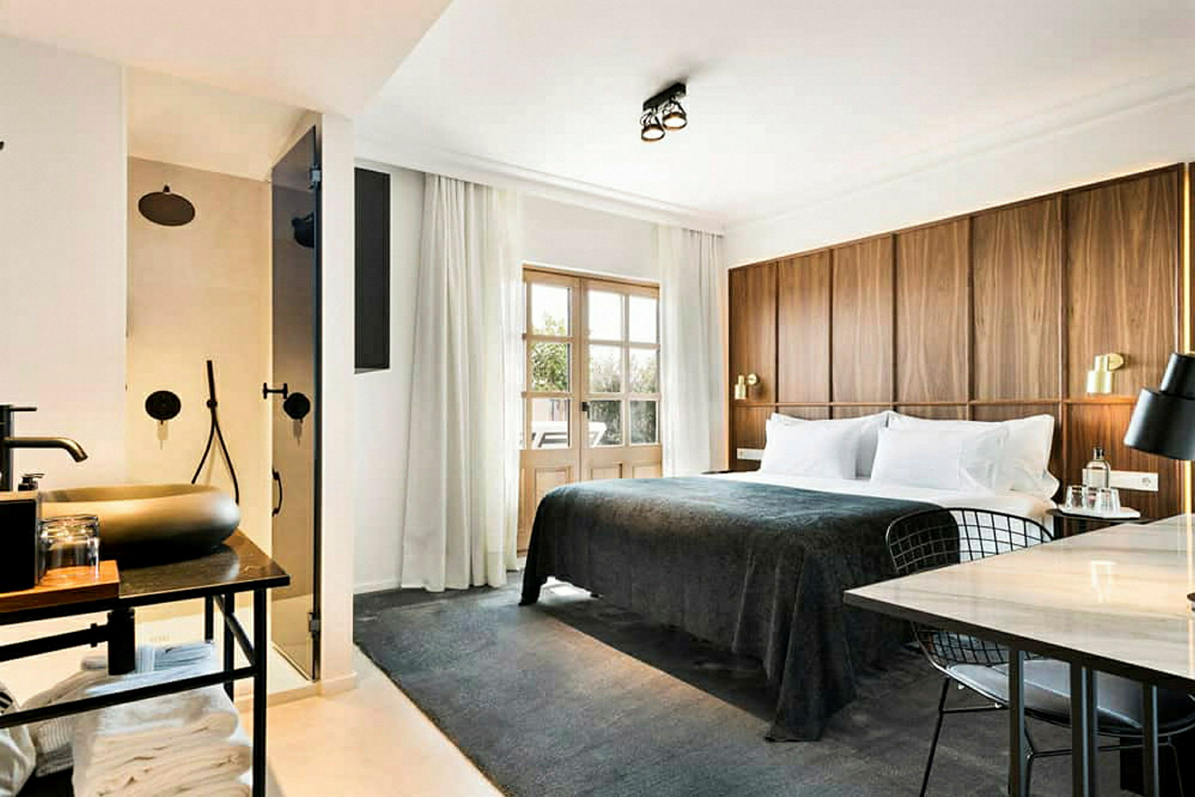 Ett hotellrum med en stor säng klädd i mörkgrått sängöverkast, en träpanel som sänggavel, en arbetsplats med bord och stol, samt ett synligt badrum med dusch bakom en glasdörr. (AI-genererad text)