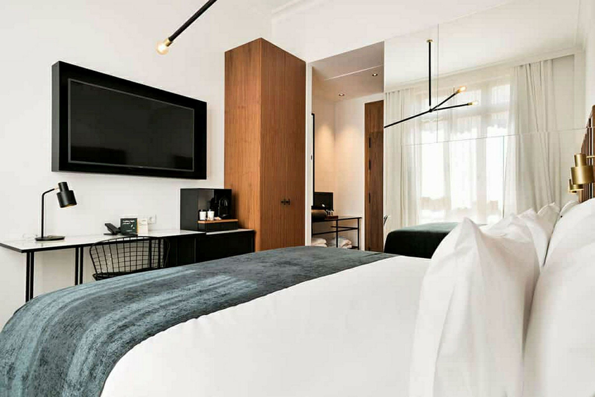 Bild av ett modernt hotellrum med en stor TV på väggen, skrivbord med lampa, en dubbelsäng med vita sängkläder och ett täcke i mörka färger. Rummet har även en garderob och stora fönster med ljusa gardiner. (AI-genererad text)