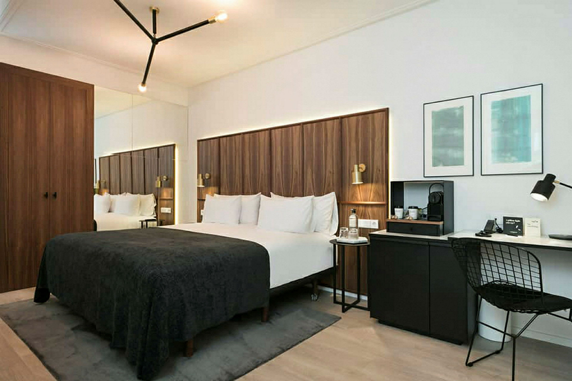 Ett modernt hotellrum med en dubbelsäng täckt av mörkt sängöverkast, träpaneler som huvudgavel, en skrivbordshörna med stol och väggkonst, samt en garderob med spegel. (AI-genererad text)