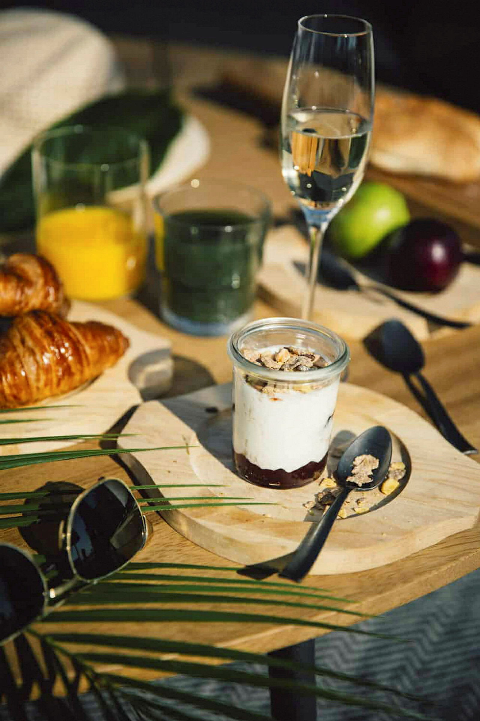 Ett frukostbord med glas av drycker, en croissant, en tallrik med yoghurt och granola samt andra detaljer som glasögon och frukter. (AI-genererad text)
