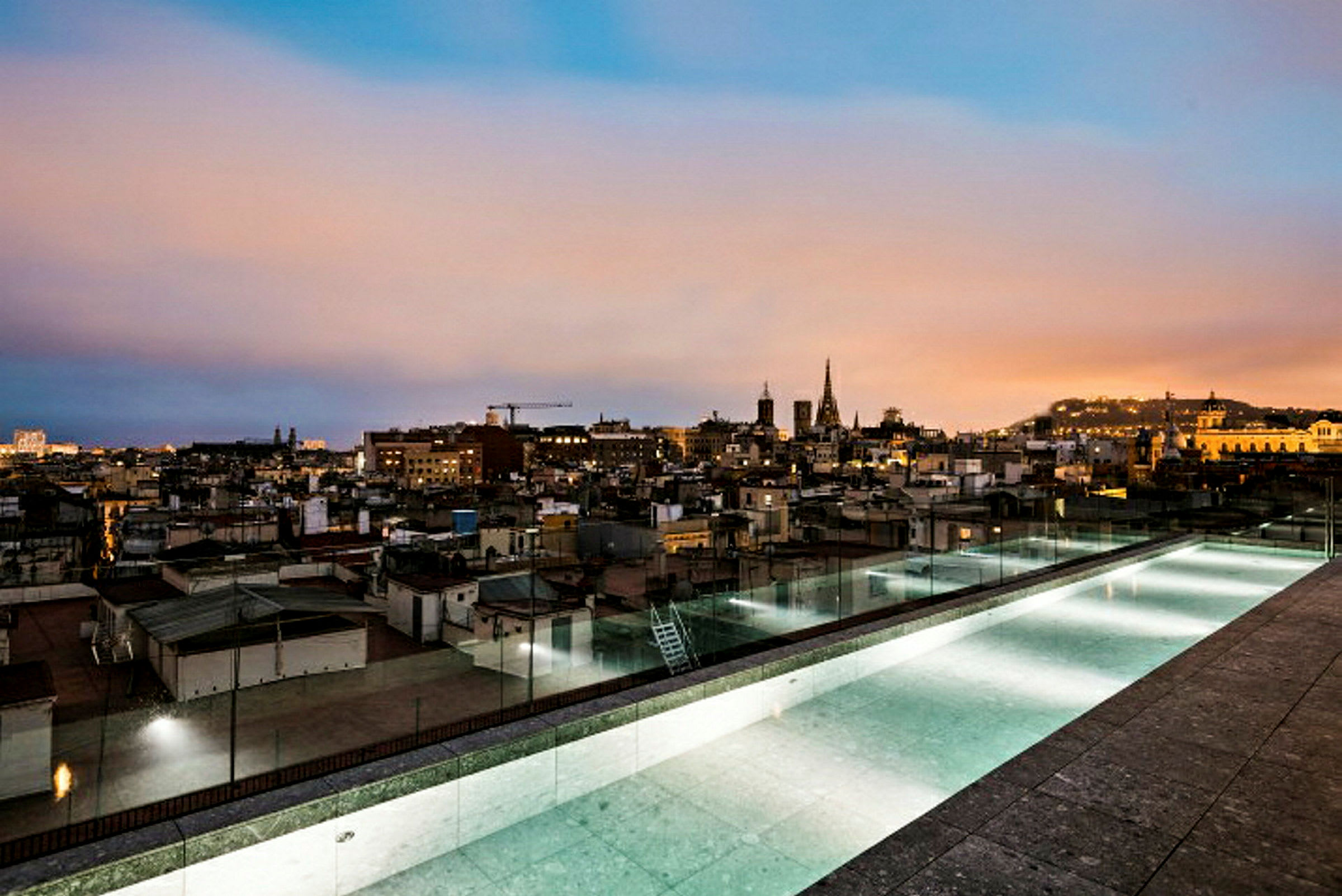 Utsikt från en takterrass med en lång pool över en stad i skymningen. (AI-genererad text)