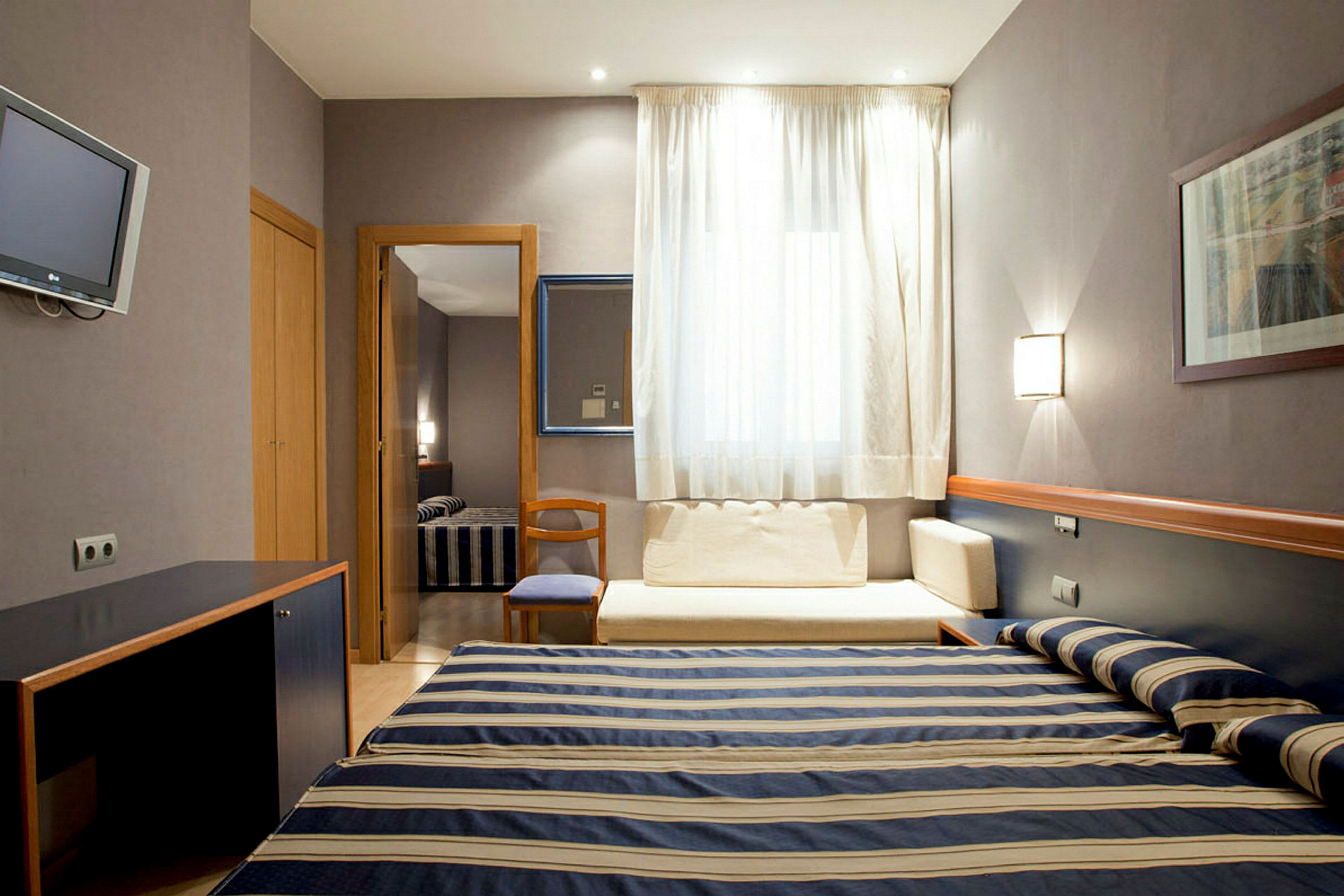 Ett hotellrum med en bäddad enkelsäng med randigt överkast, ett skrivbord, och en sittplats vid ett fönster med ljusa gardiner. (AI-genererad text)