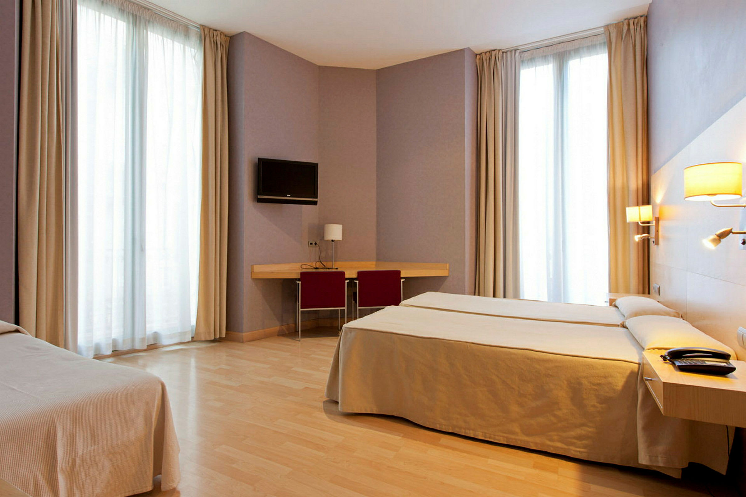 Ett hotellrum med två sängar, skrivbord med stol, väggmonterad TV och stora fönster med gardiner. (AI-genererad text)