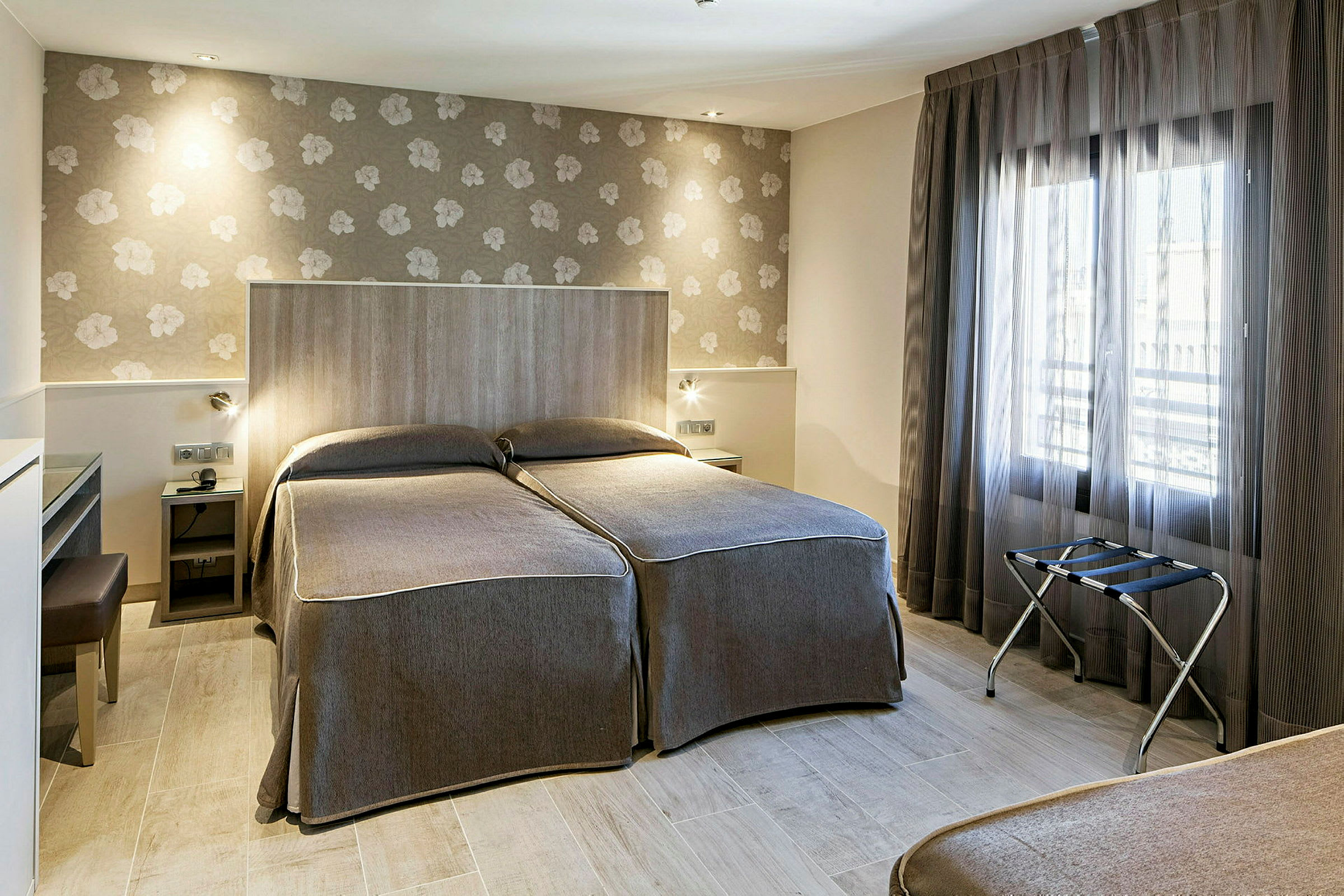 Ett hotellrum med två enkelsängar, tapetserade väggar och ett stort fönster med genomskinliga gardiner. (AI-genererad text)
