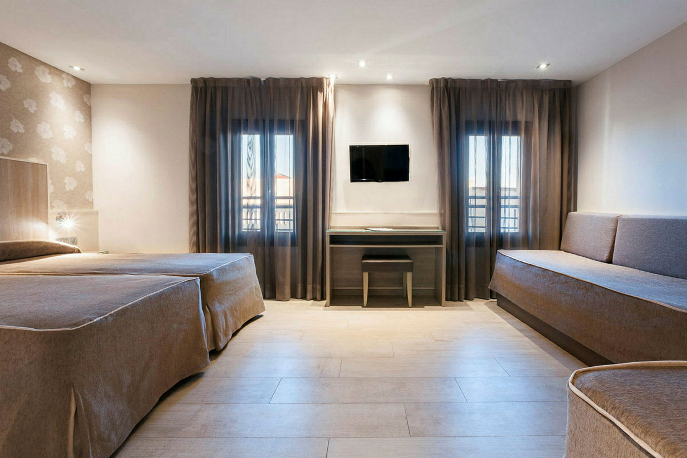 Ett hotellrum med två sängar, en soffa, två fönster med mörka gardiner och en väggmonterad TV ovanför ett skrivbord. (AI-genererad text)