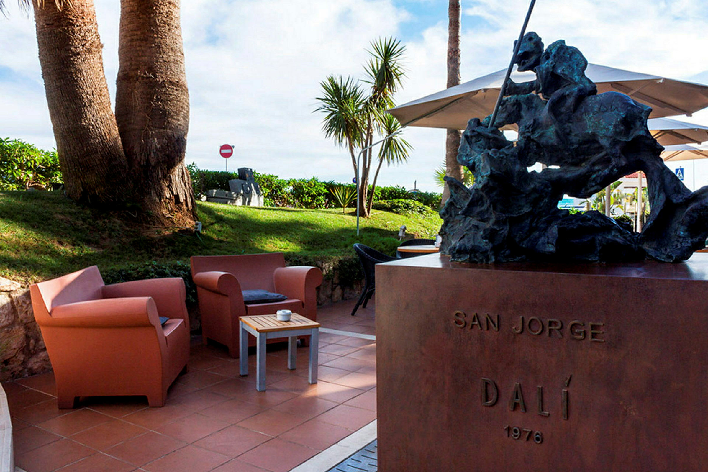 En staty står på en piedestal, med texten 'SAN JORGE DALÍ 1976'. I bakgrunden syns en gräsmatta med palmer och sittmöbler på en terrass. (AI-genererad text)