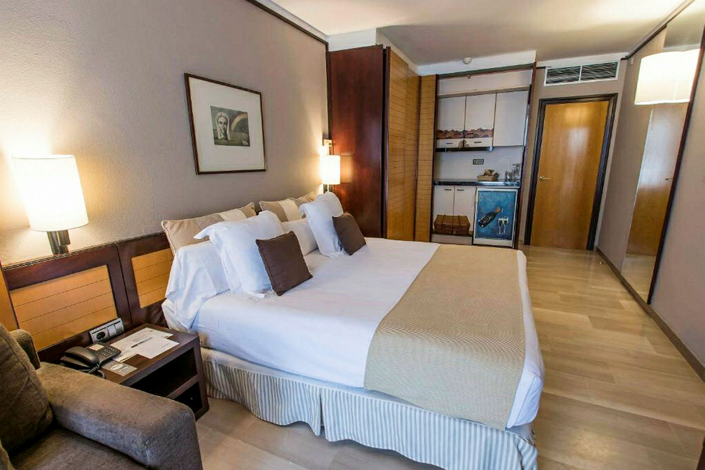 Ett hotellrum med en stor säng, kuddar, sängbord och en liten köksdel med skåp. (AI-genererad text)