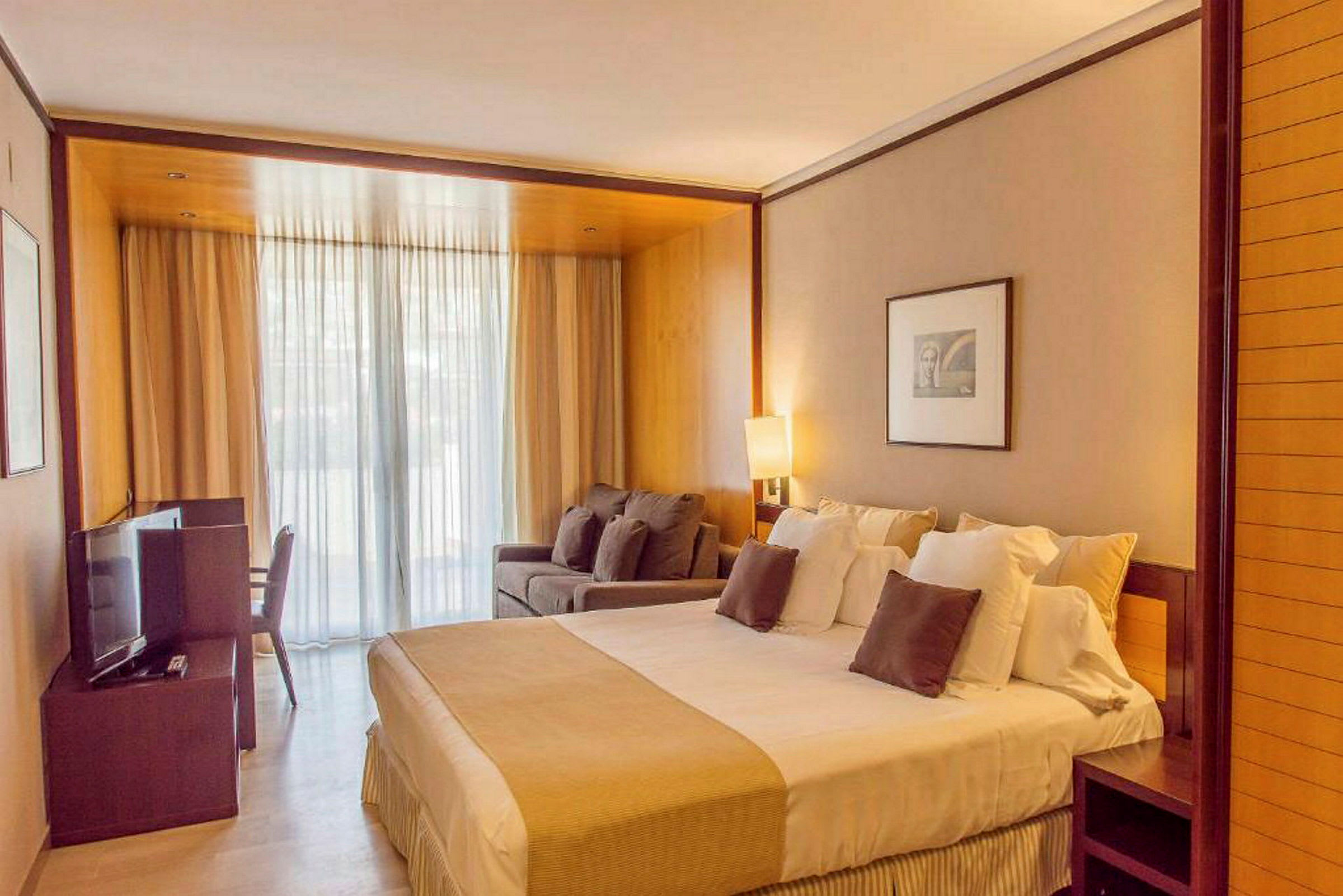 Ett hotellrum med en stor säng, soffa och TV. Rummet har också en glasdörr med gardiner som leder till en balkong. (AI-genererad text)