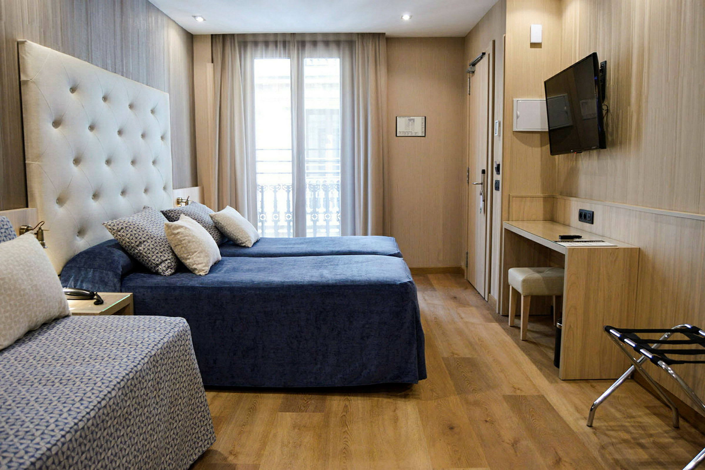 Ett hotellrum med två sängar klädda i blåa sängöverkast och flera kuddar. Rummet har en ljus inredning med trädetaljer, ett skrivbord, en stol och en TV monterad på väggen. (AI-genererad text)