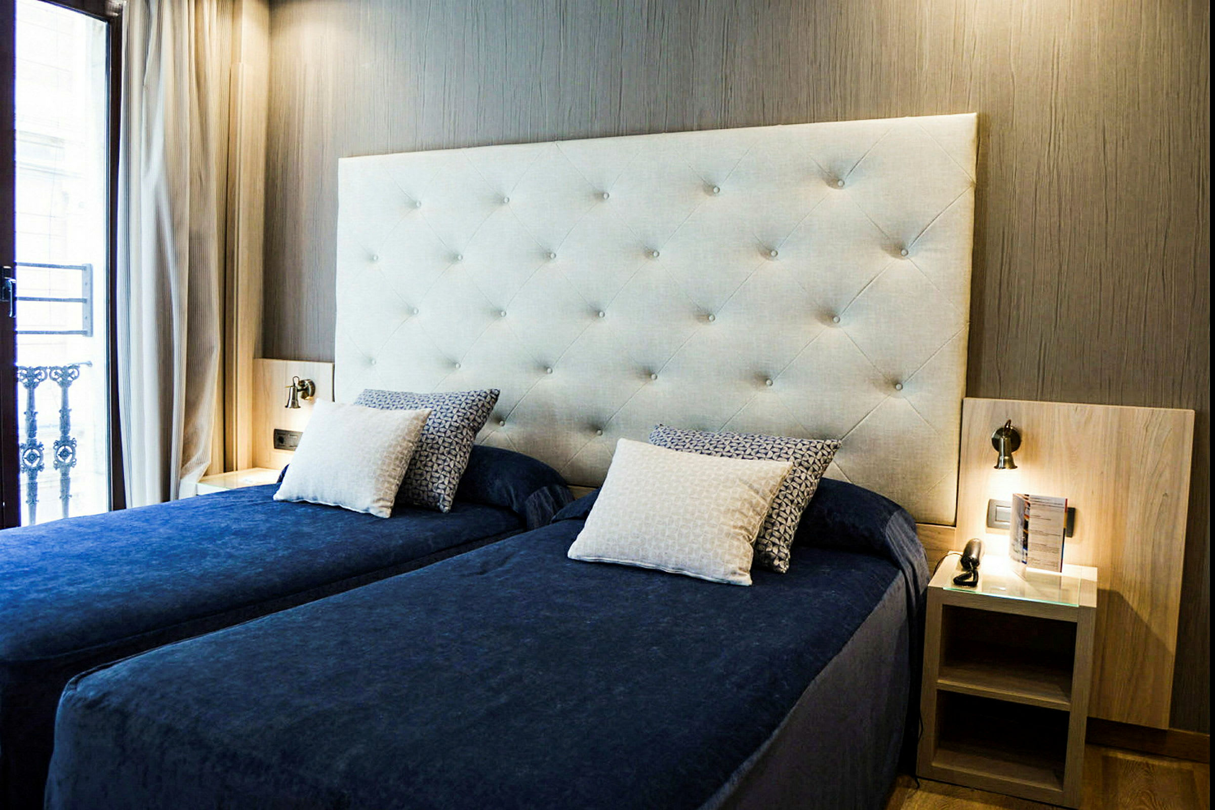 Ett hotellrum med två enkelsängar klädda i blått och en vadderad sänggavel, bredvid varsitt nattduksbord. (AI-genererad text)