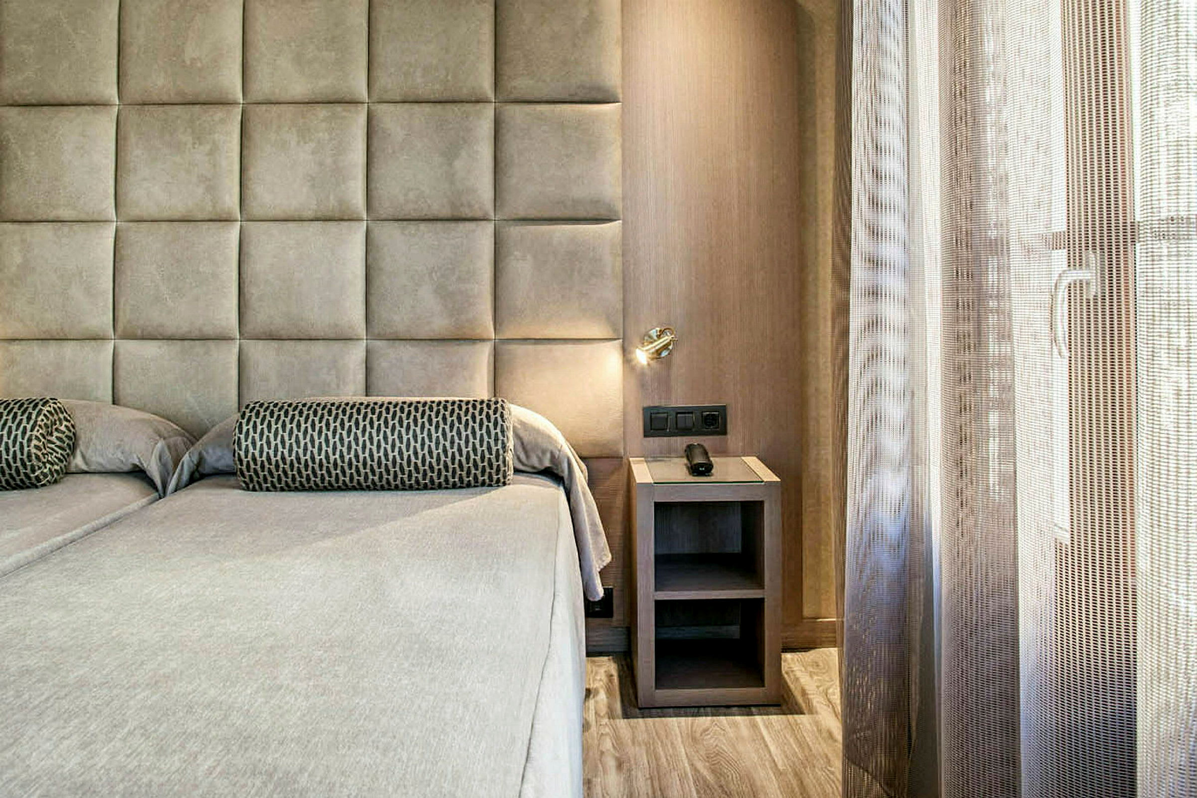 Ett hotellrum med två sängar intill en mjukt vadderad vägg och ett nattduksbord med inbyggd lampa. Ett ljusstrilande fönster täcks delvis av ett nätliknande draperi. (AI-genererad text)