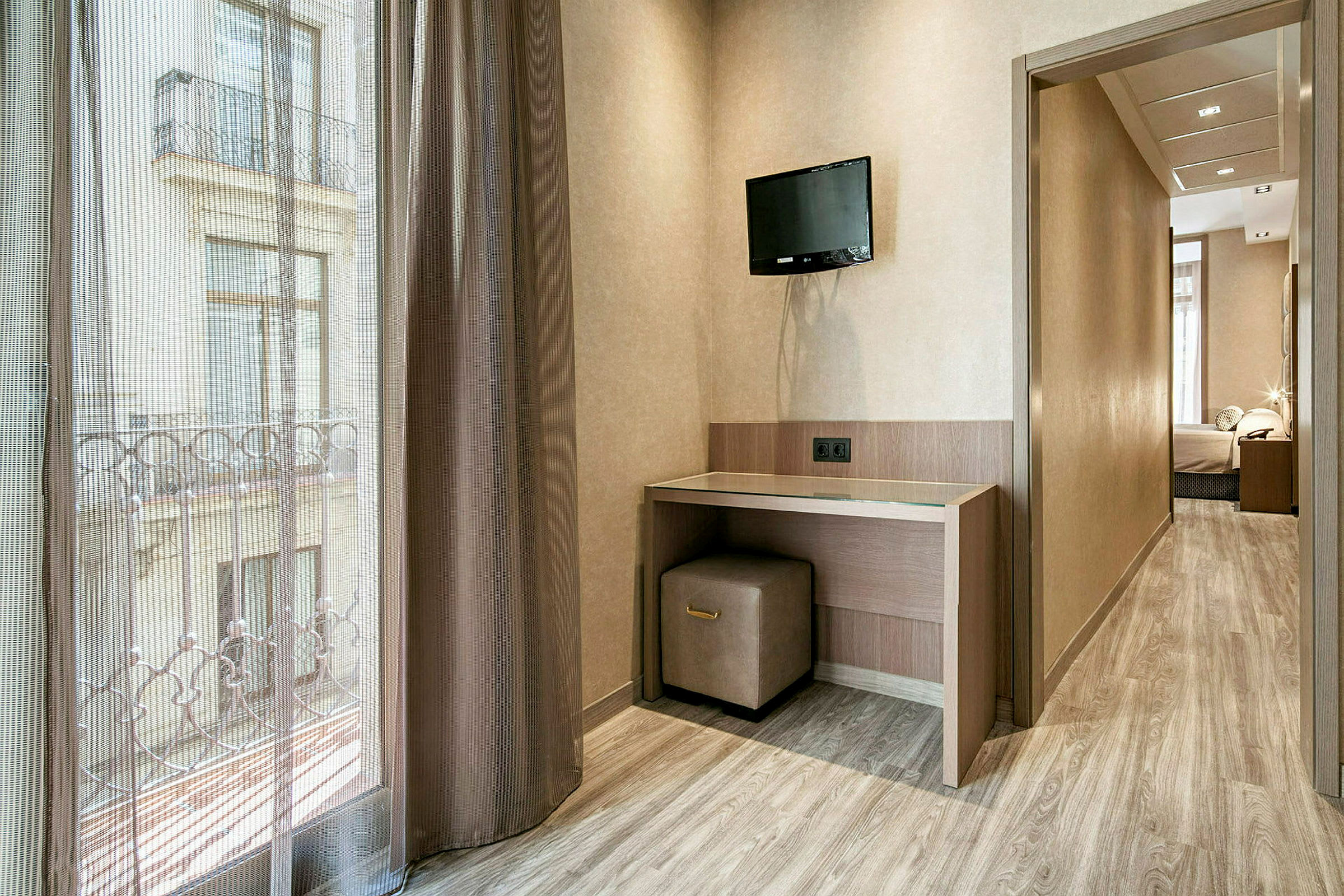 Ett hotellrum med en liten skrivbordshörna. En TV är monterad på väggen ovanför skrivbordet, och ett fönster med gardiner ger utsikt över omgivningen. (AI-genererad text)