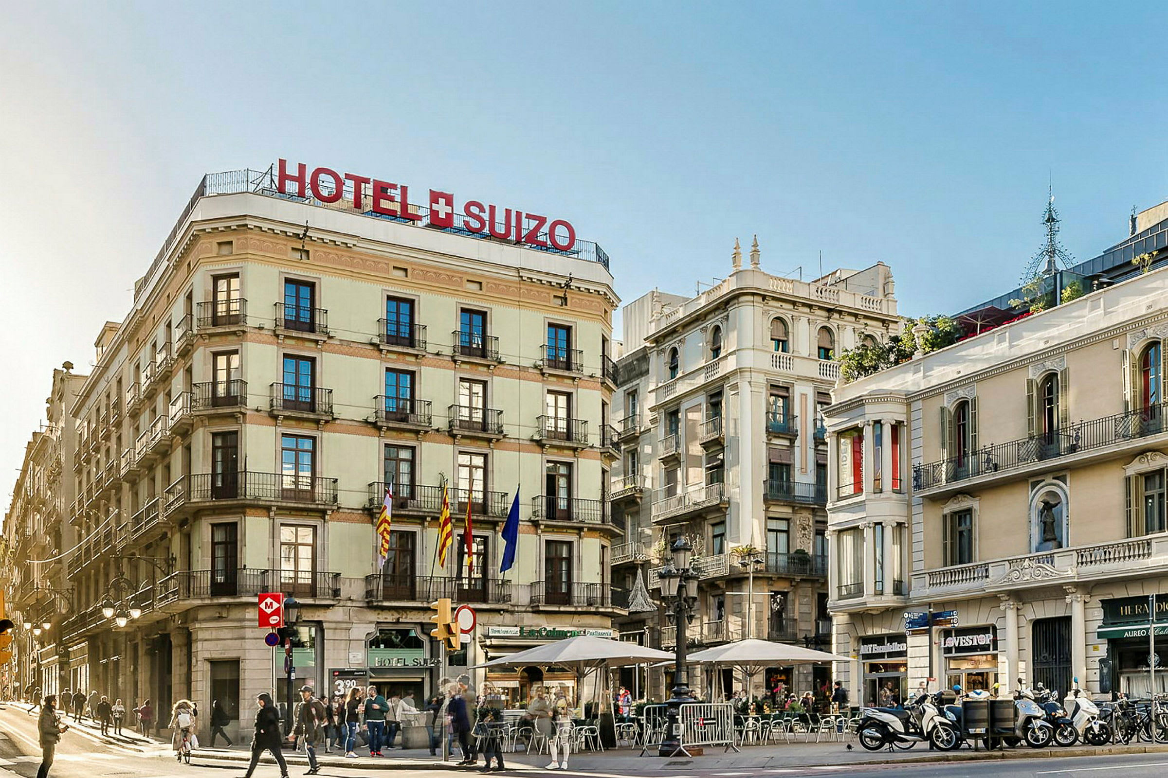Ett hotell med namnet "Hotel Suizo" på en hörna med flera våningar och balkonger, omgiven av andra byggnader och människor som promenerar på gatan. (AI-genererad text)