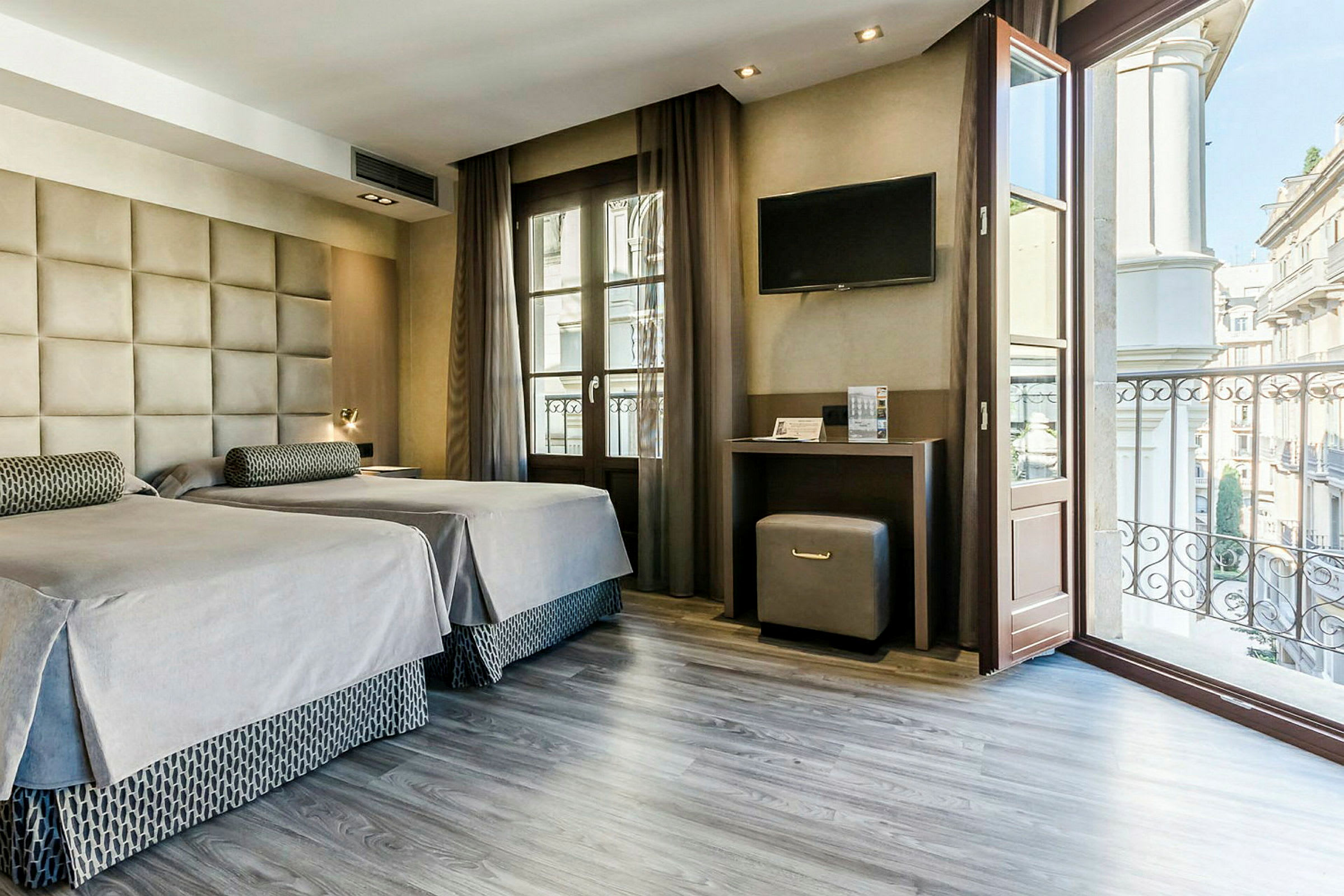 Ett hotellrum med två enkelsängar, en skrivbordsyta med TV ovanför, och ett fönster som vetter mot en balkong med utsikt över stadsmiljön. (AI-genererad text)