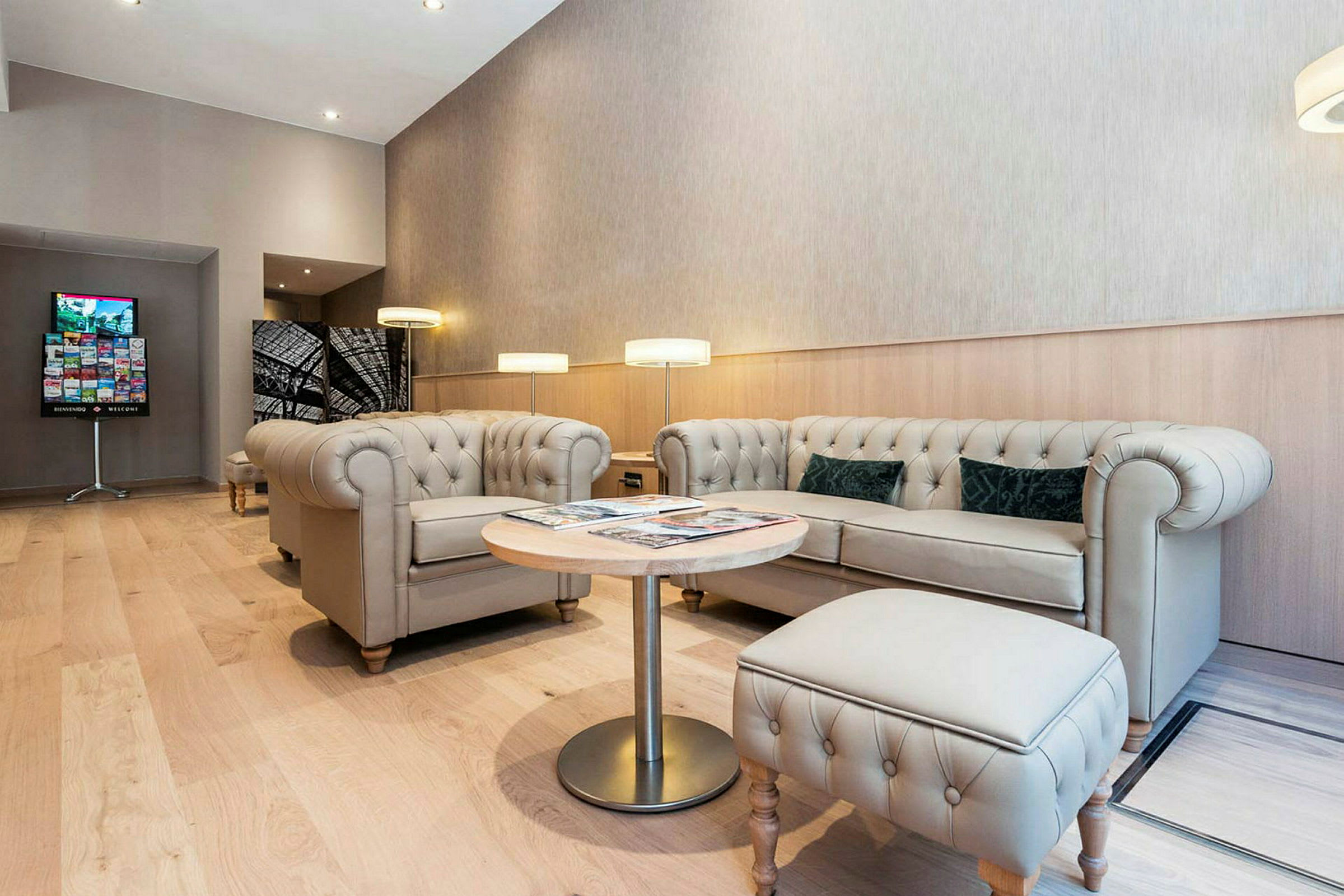 Ett modernt loungeområde med beige chesterfieldsoffor och fåtöljer, ett runt träbord, och golvlampor. Golvet är av ljus träparkett. (AI-genererad text)
