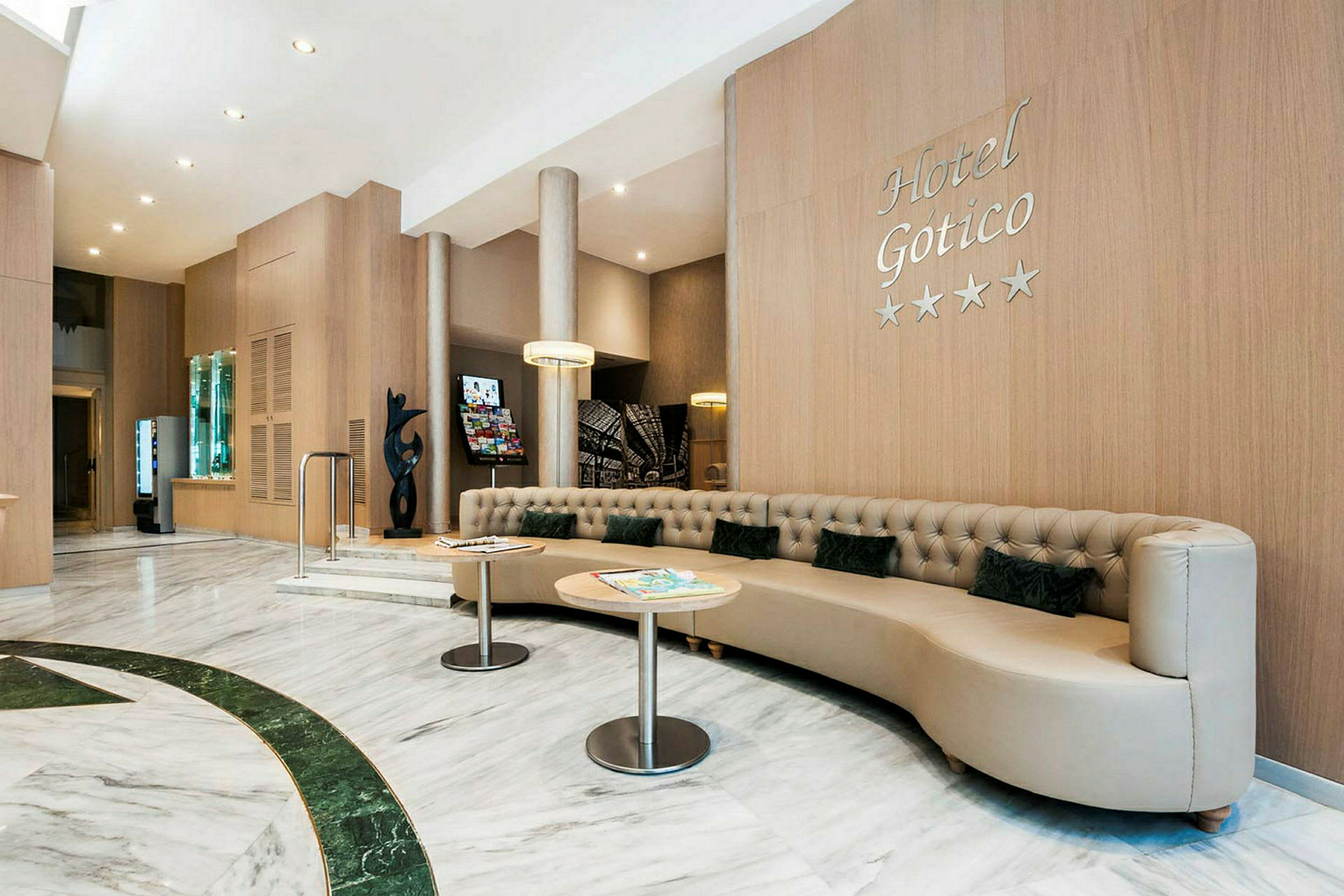 Hotellobby med modernt, ljust inredd interiör. En stor, beige soffa med små kuddar och två runda bord syns. Väggarna är klädda i träpanel och en skylt med texten "Hotel Gotico" syns. (AI-genererad text)