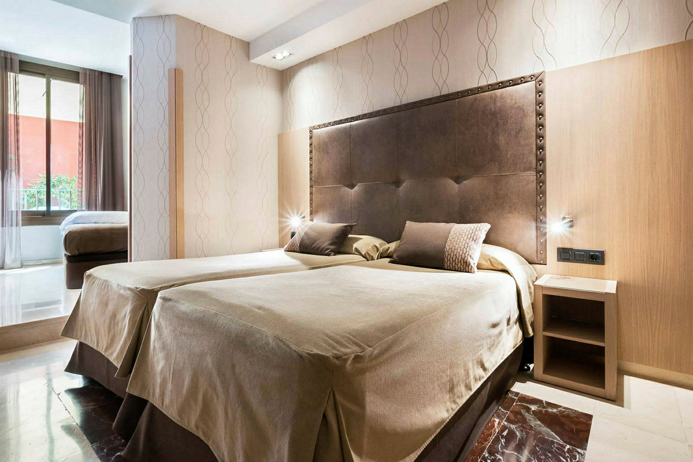Ett hotellrum med två enkelsängar bredvid varandra och en huvudsäng i mörkt material. Det finns ett litet sängbord och en stor fönsteryta som släpper in naturligt ljus. (AI-genererad text)