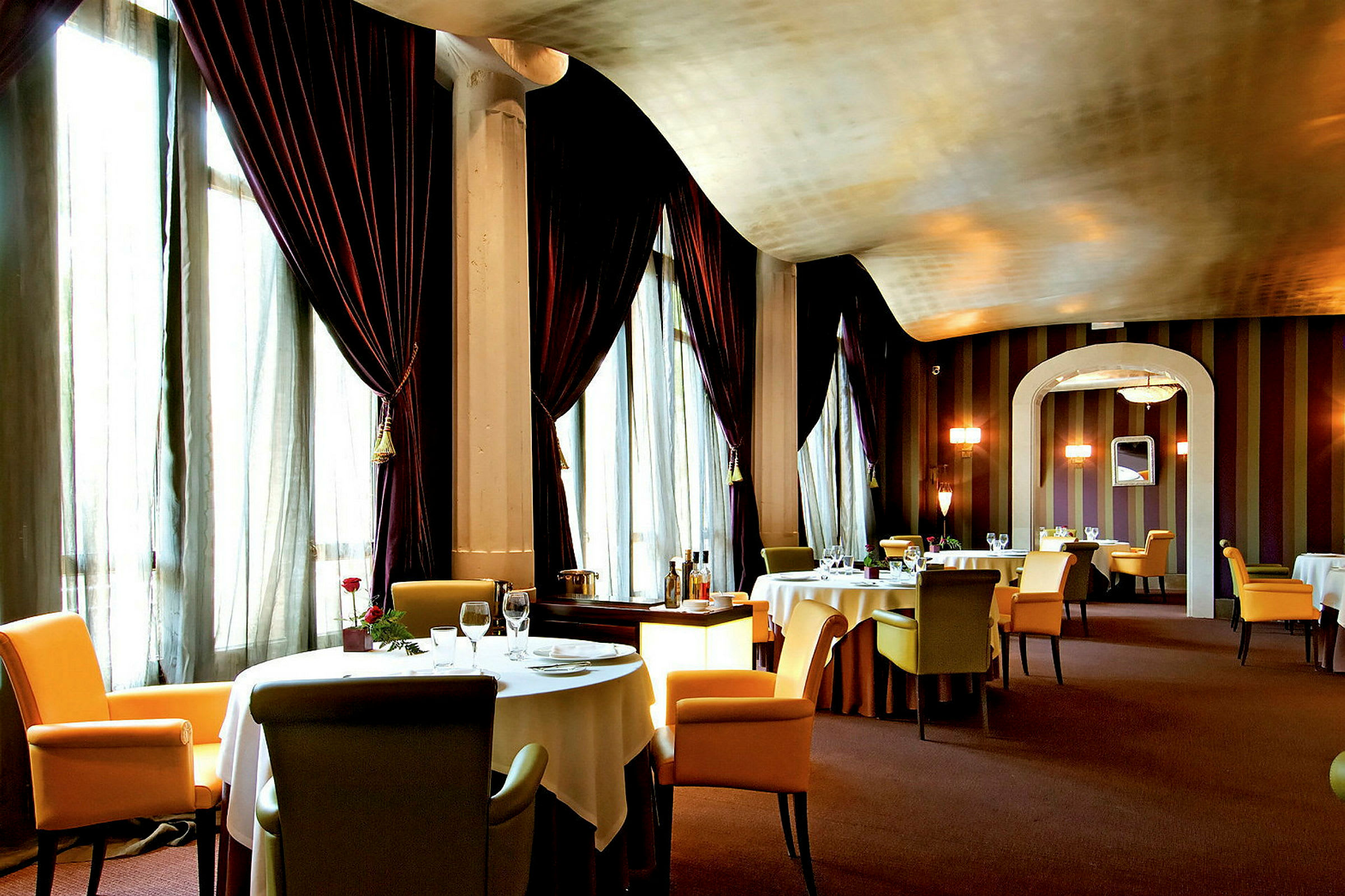 En elegant restaurang med dukade bord och bekväma stolar, omgiven av stora gardiner och dämpad belysning. (AI-genererad text)