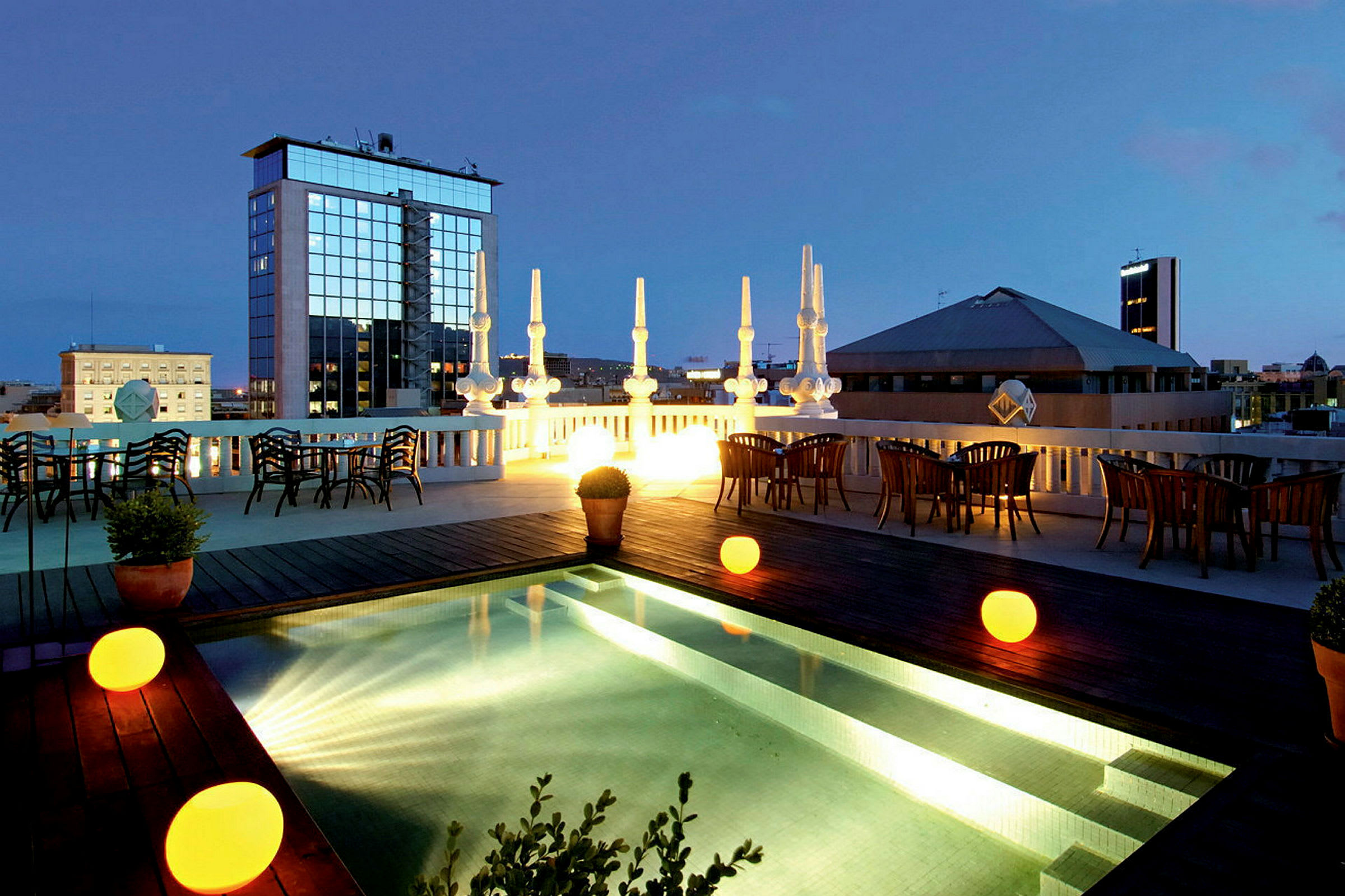 En takterrass med en upplyst pool, omgivande bord och stolar samt stadsvy i bakgrunden under skymningen. (AI-genererad text)