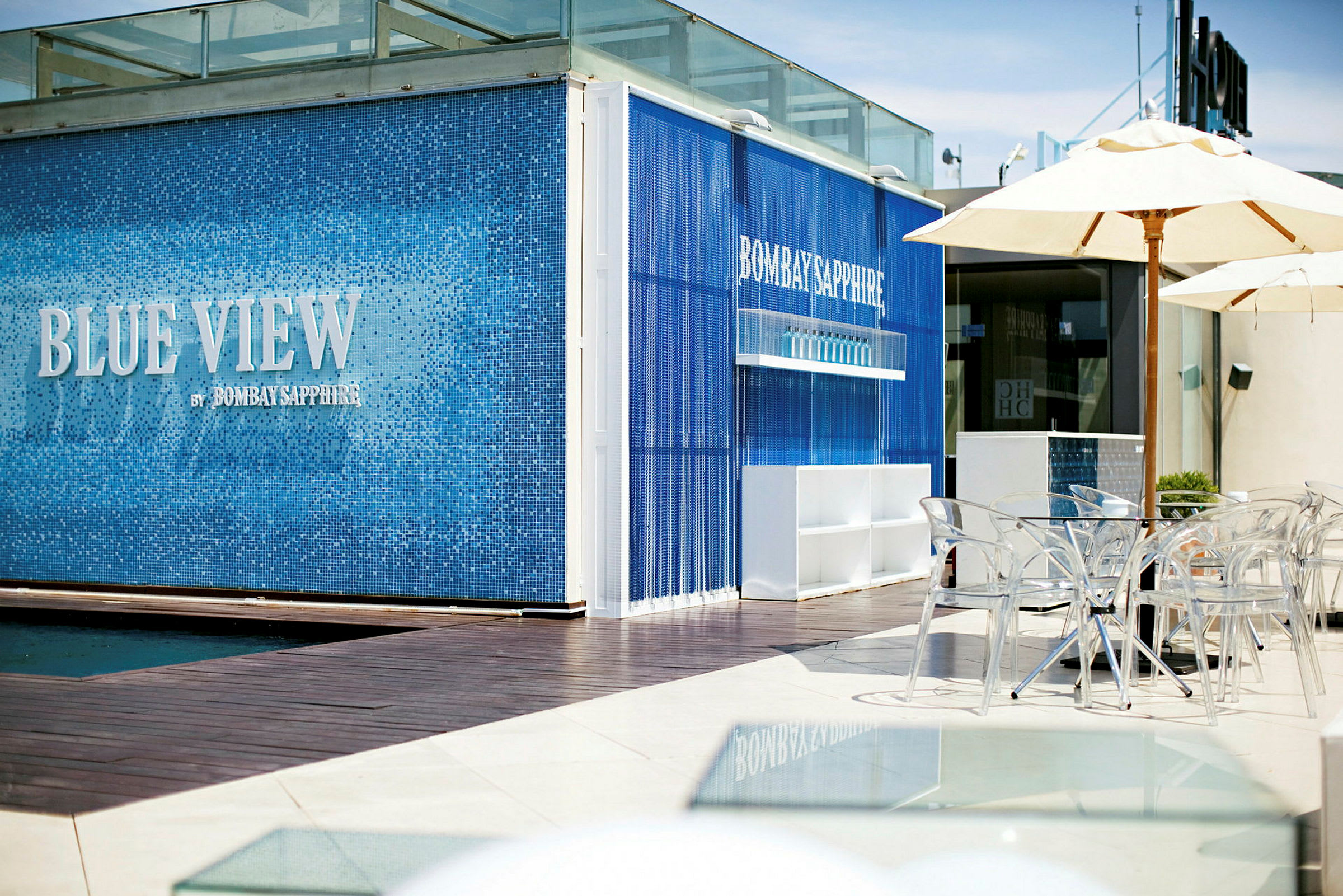 En modern bar exteriör i blått kakel med texten "Blue View by Bombay Sapphire". Framför finns genomskinliga stolar och parasoller. (AI-genererad text)
