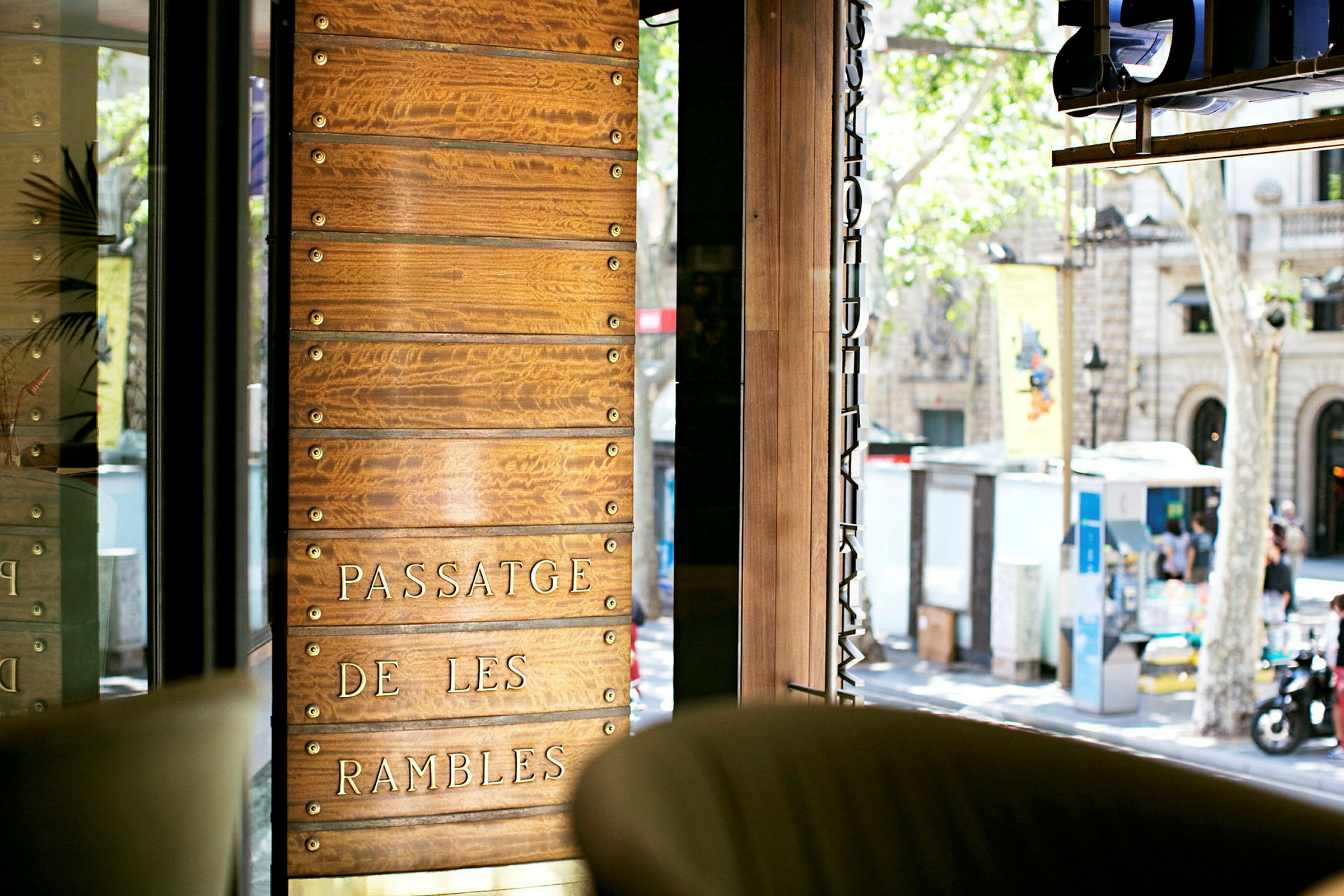 Träpanel med texten "Passatge de les Rambles" sedd från en inomhusmiljö med utsikt över gatan. (AI-genererad text)