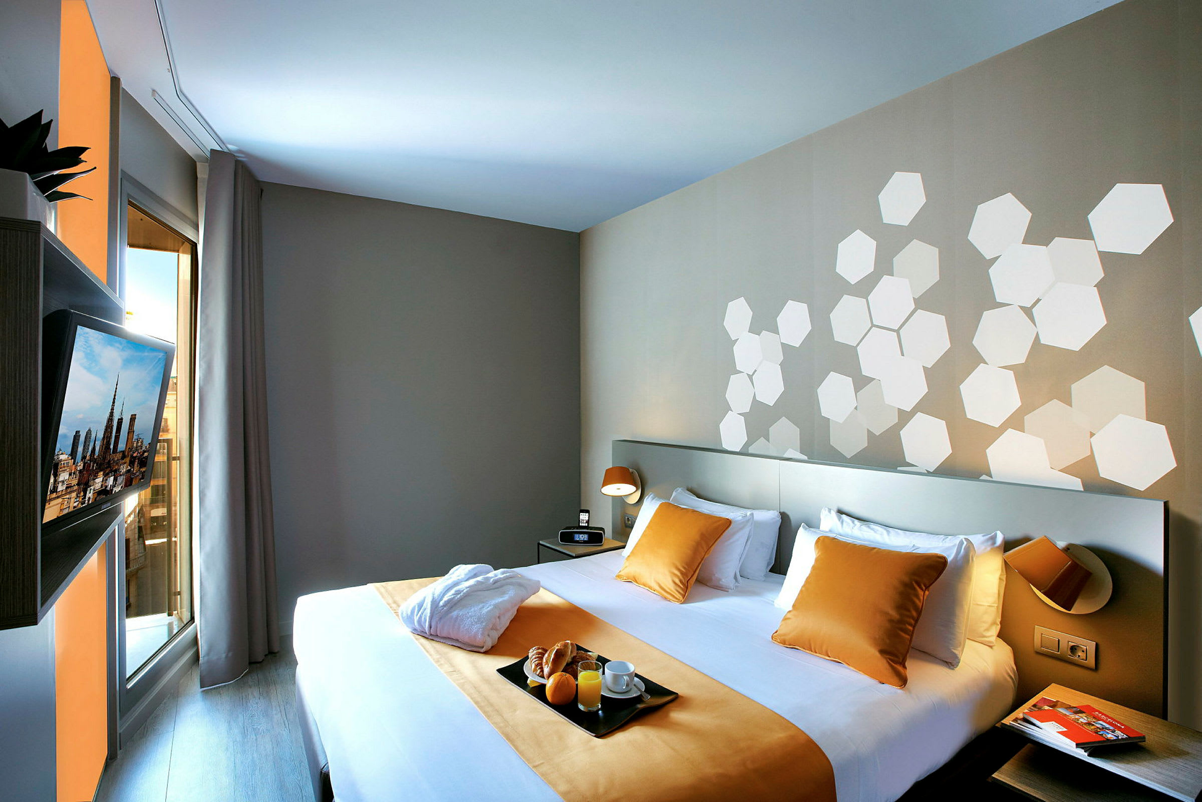 Ett hotellrum med en dubbelsäng som är bäddad med vita lakan och gula kuddar och överkast. På väggen finns hexagonala mönster i vitt. Ett fönster med gardiner och en väggmonterad TV syns också. (AI-genererad text)