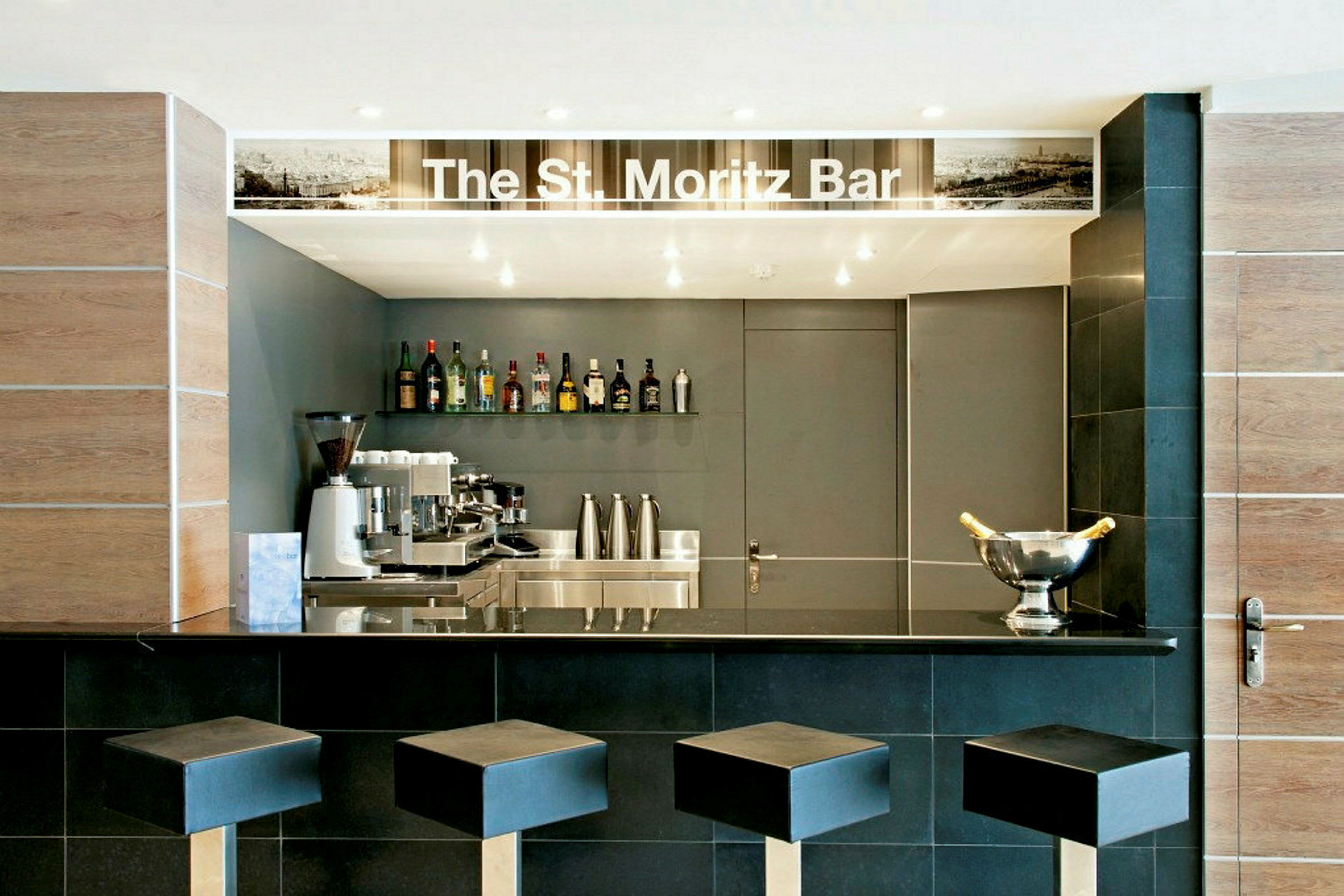 En bar med namnet 'The St. Moritz Bar' som har svarta stolar och en välutrustad disk med flaskor, kaffemaskin och champagnekyl. (AI-genererad text)