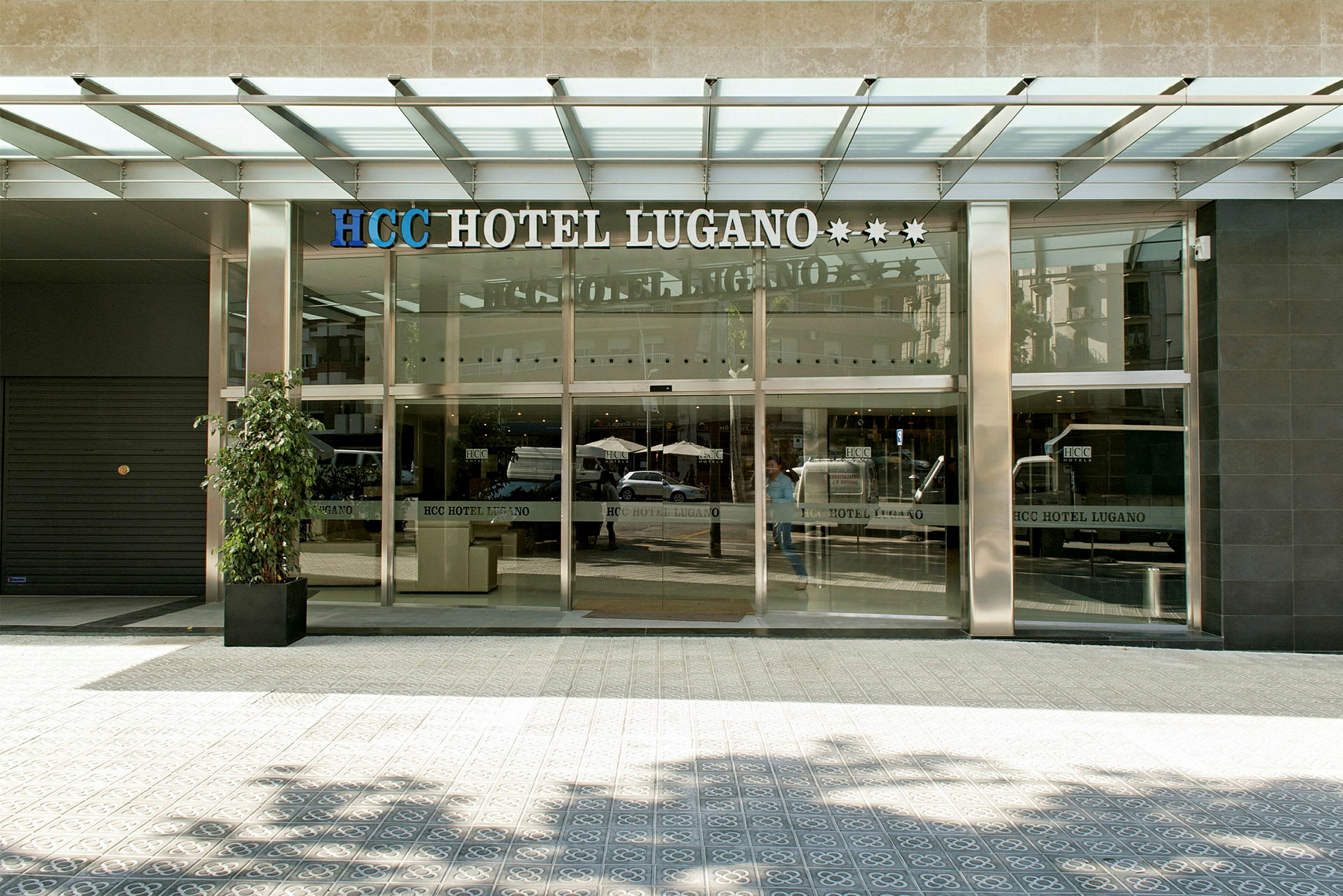 Entrén till HCC Hotel Lugano med glasdörrar och en modern byggnadsfasad. (AI-genererad text)