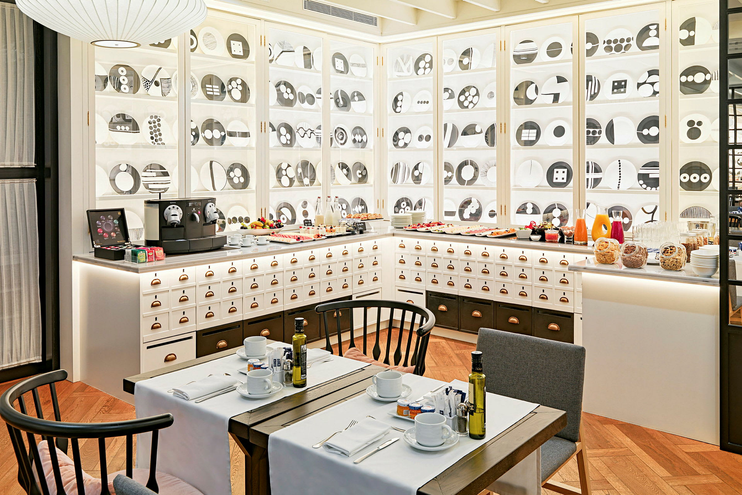 Ett café eller restaurang med vita skåp dekorerade med tallrikar i ett geometriskt mönster, en serveringsstation med olika drycker och mat samt dukade bord med stolar i förgrunden. (AI-genererad text)