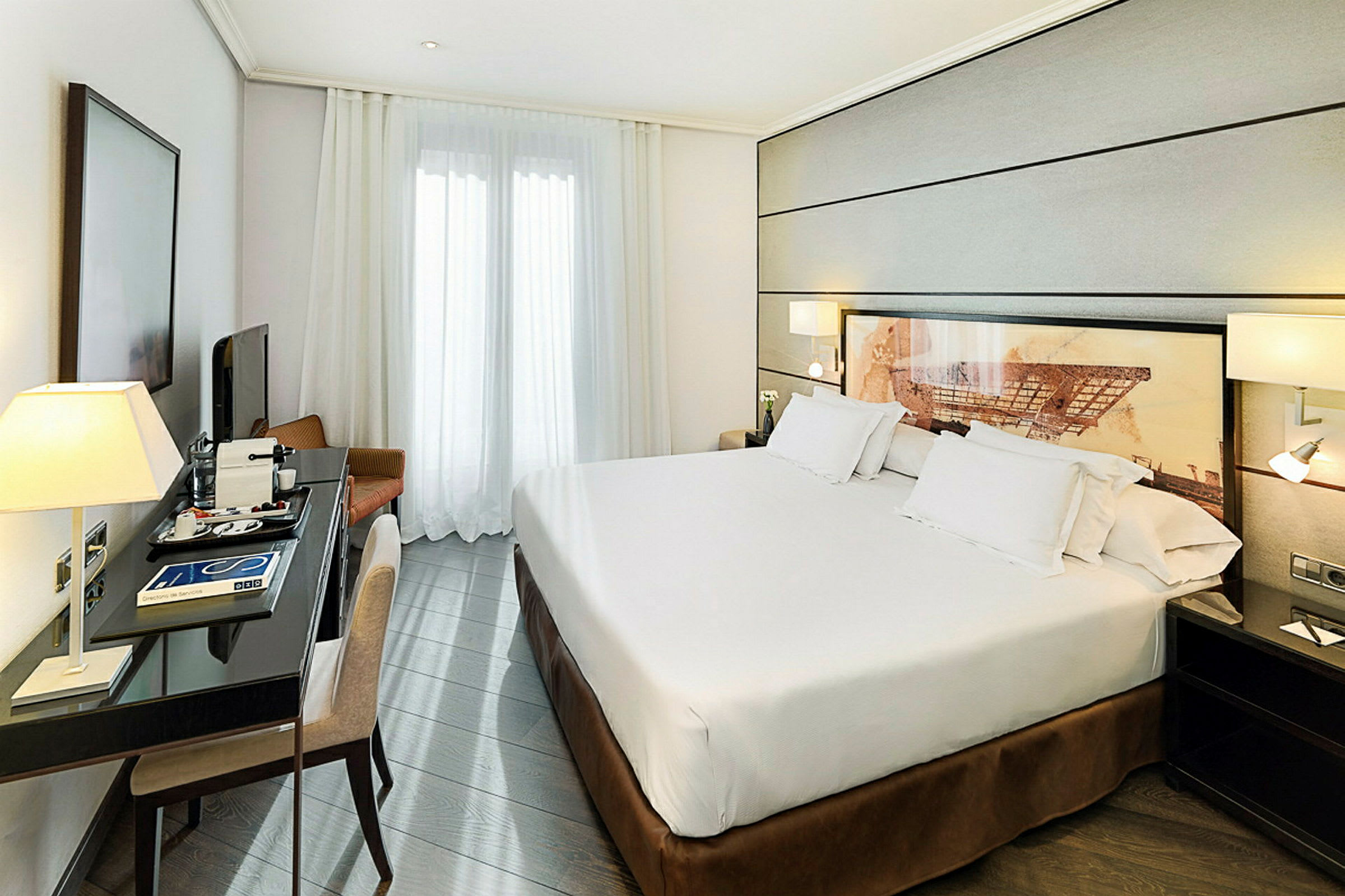 Ett hotellrum med en dubbelsäng, ett skrivbord med lampa och stol samt en TV. Ett stort fönster täckt av vita gardiner syns i bakgrunden. (AI-genererad text)