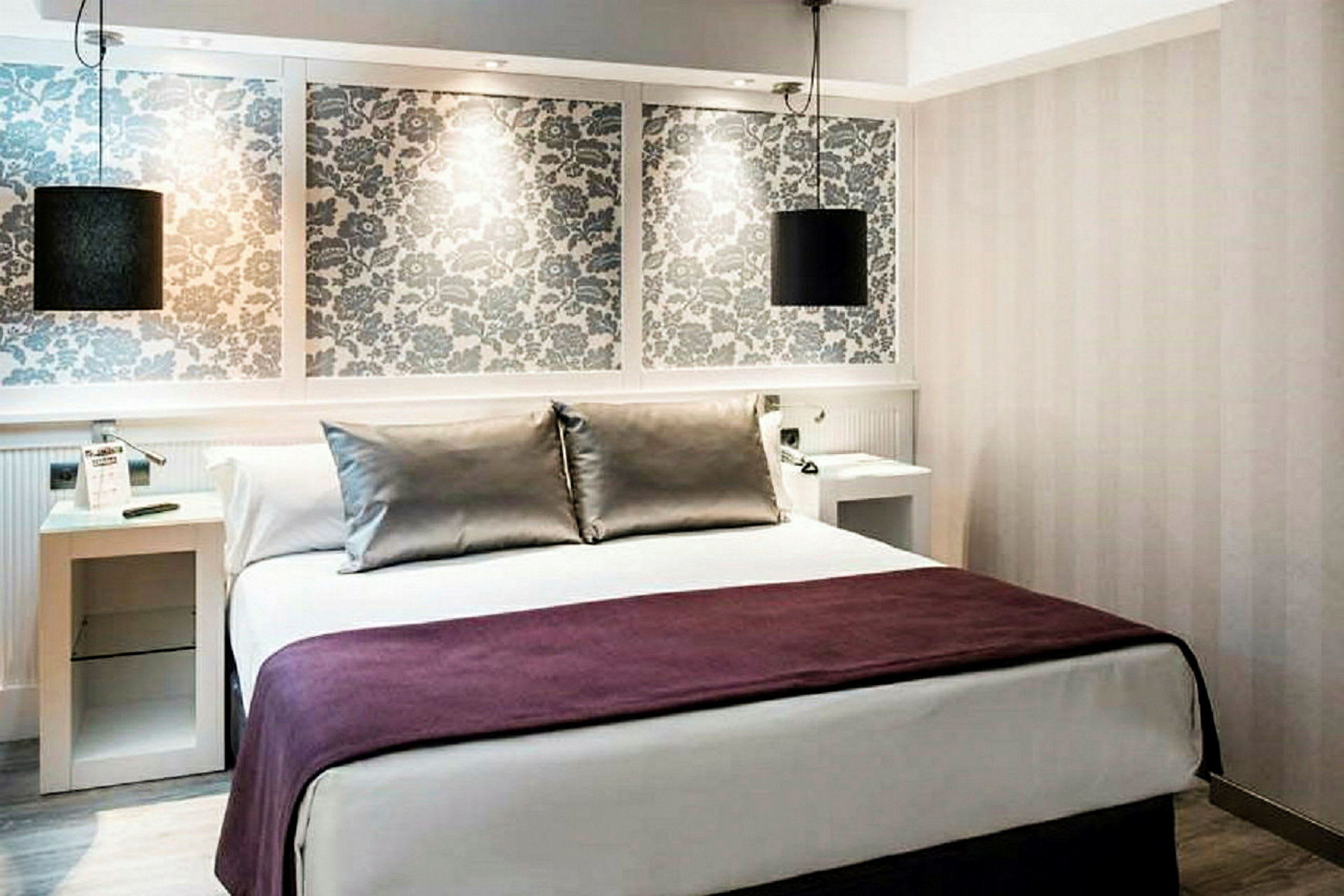 Ett hotellrum med en dubbelsäng klädd i vitt och mörkrött överkast. Bakom sängen finns en dekorativ vägg med blommigt mönster samt hängande lampor. (AI-genererad text)