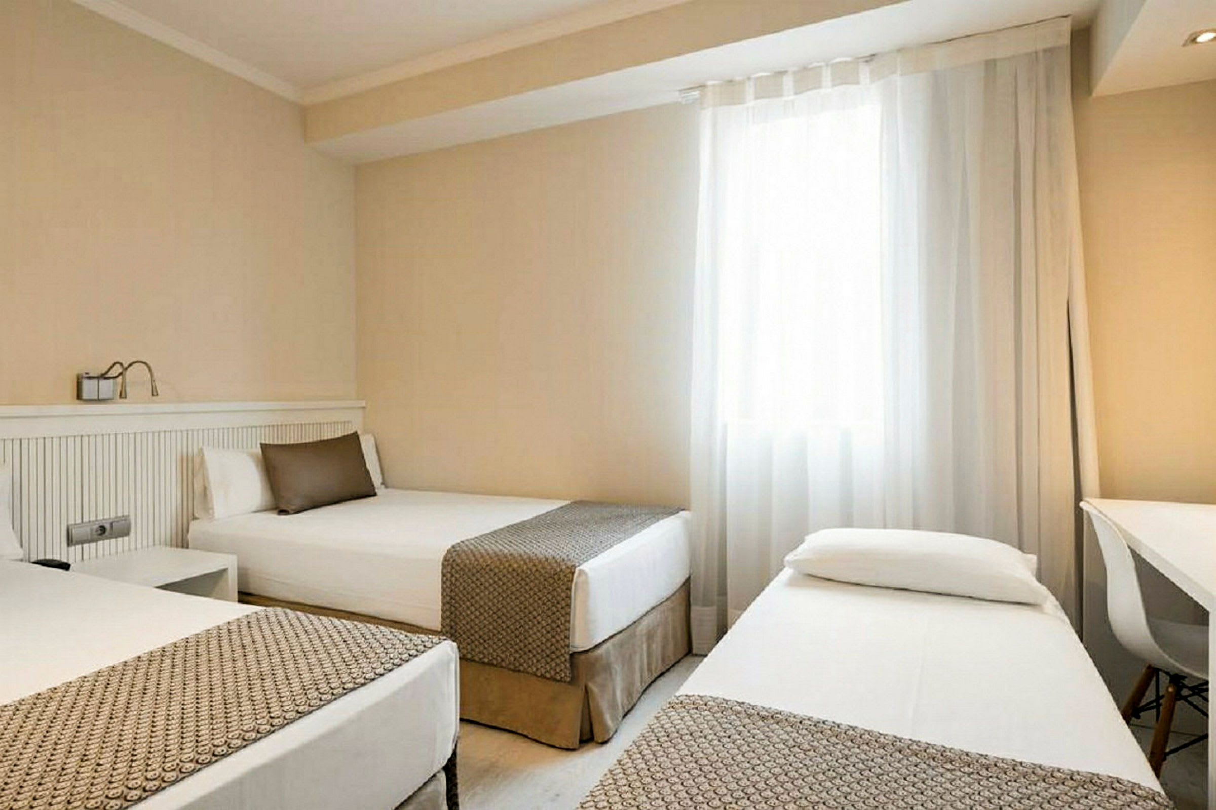 Ett hotellrum med tre enkelsängar och ljus inredning, inklusive ett fönster med genomsläppliga draperier. (AI-genererad text)