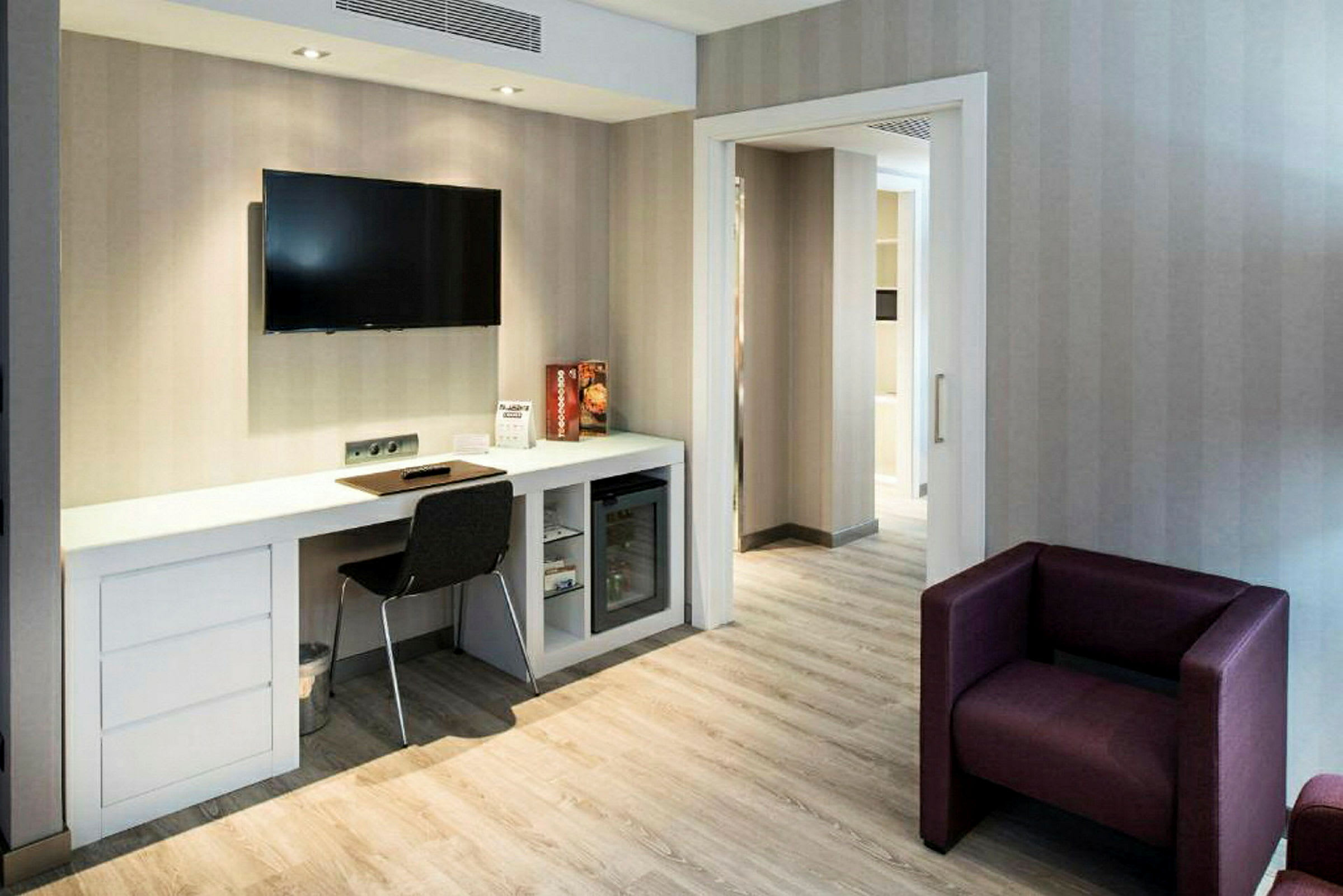 Ett hotellrum med ett skrivbord, en stol och en väggmonterad TV. Ett lila lounge-set syns i förgrunden. (AI-genererad text)
