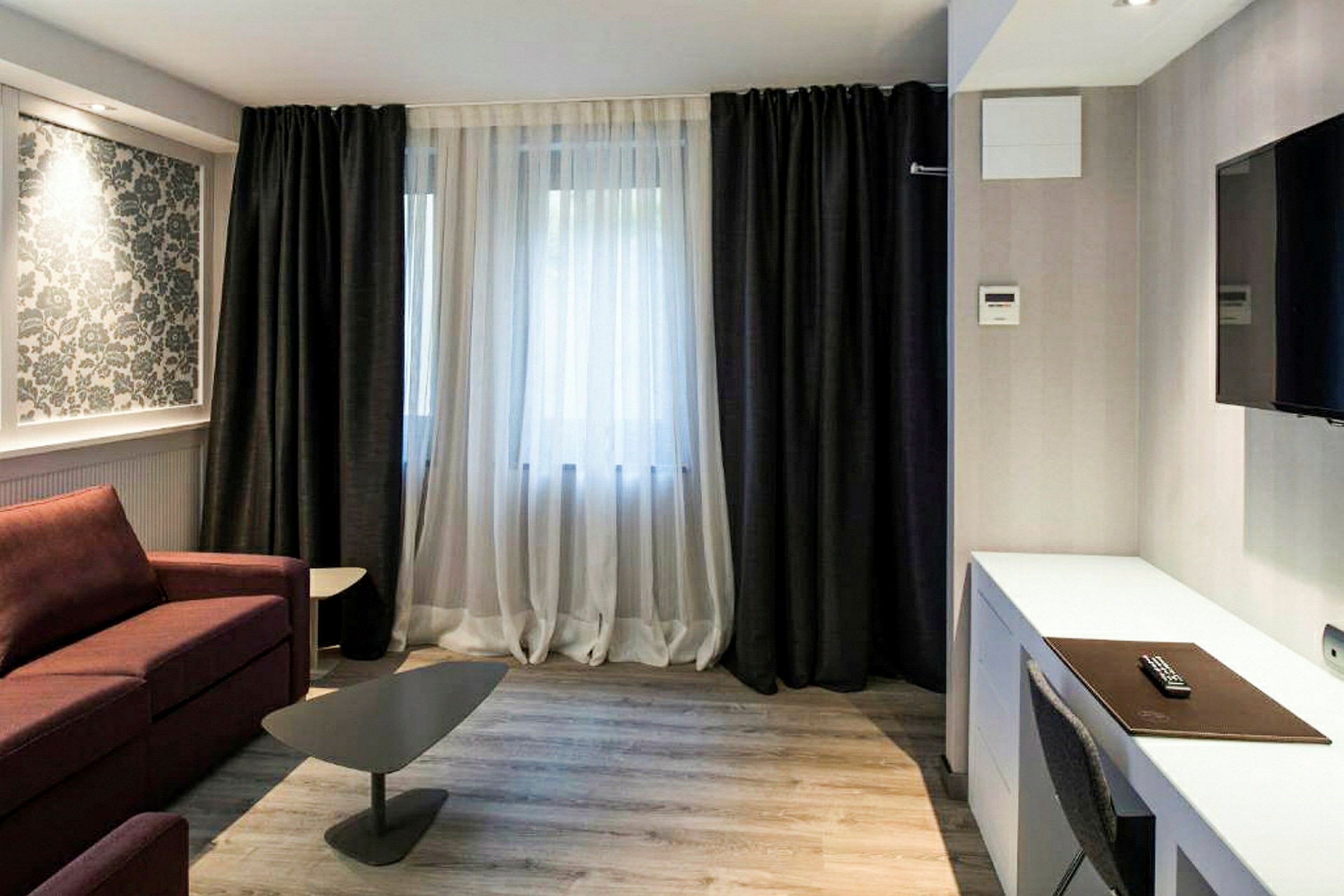 Ett modernt inrett hotellrum med mörka gardiner, ett skrivbord, en TV på väggen och en soffa med ett litet bord. (AI-genererad text)