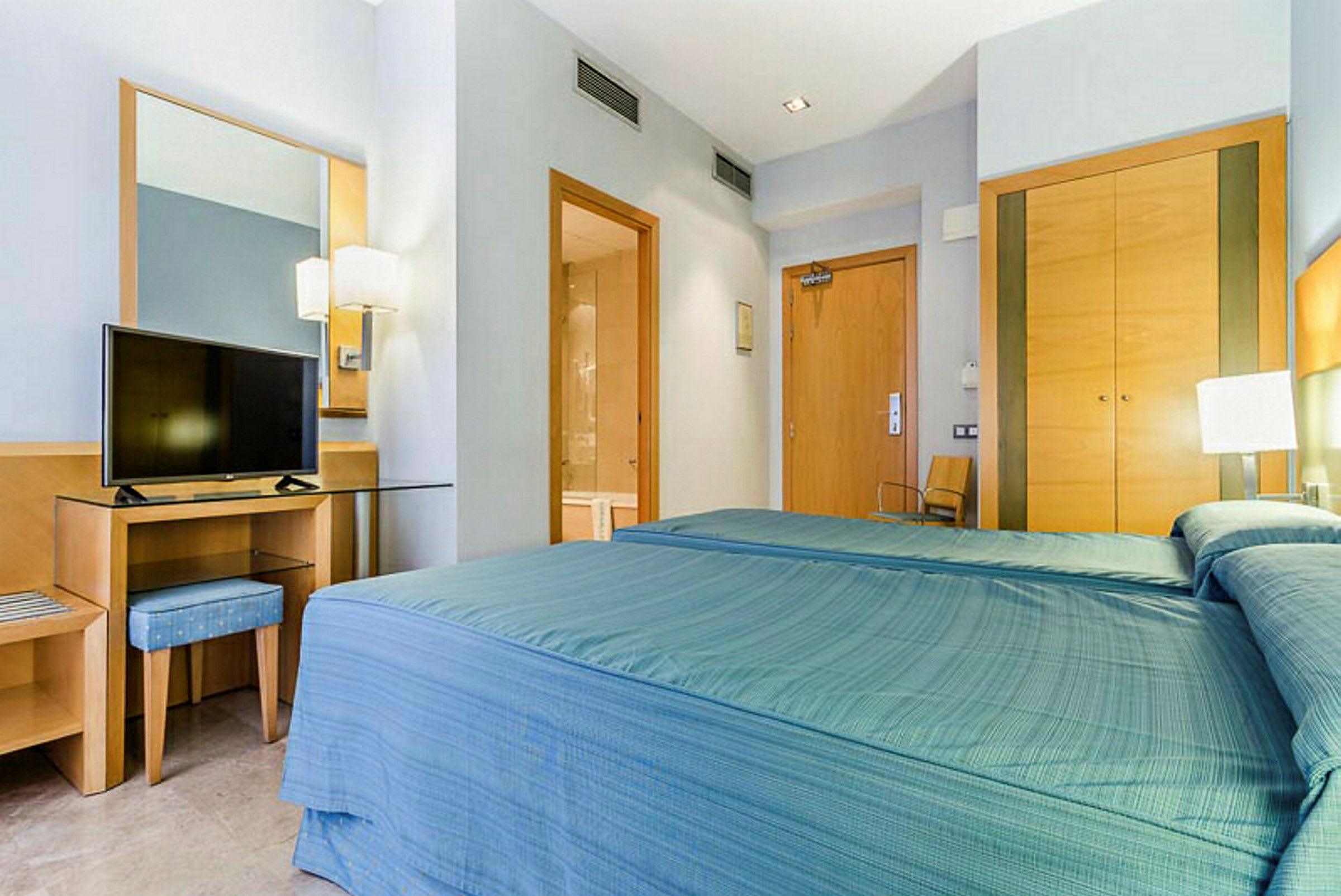 Ett hotellrum med en bäddad säng med blått överkast, ett skrivbord med en spegel och en TV ovanpå, samt garderober, badrumsdörr och ytterdörr. (AI-genererad text)