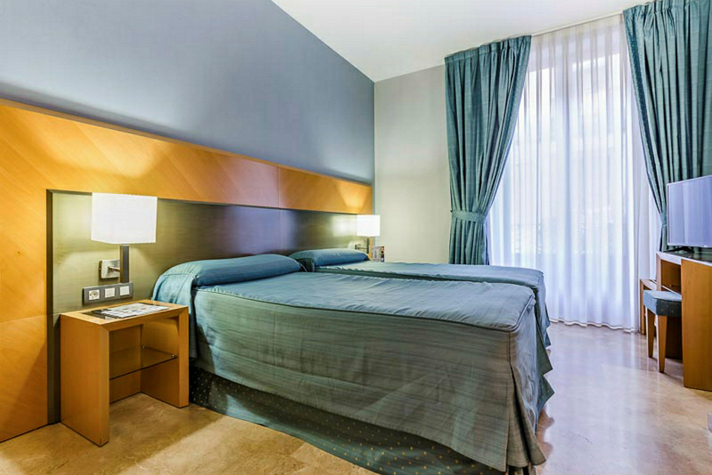Ett hotellrum med två enkelsängar, sängbord, gardiner och TV. (AI-genererad text)
