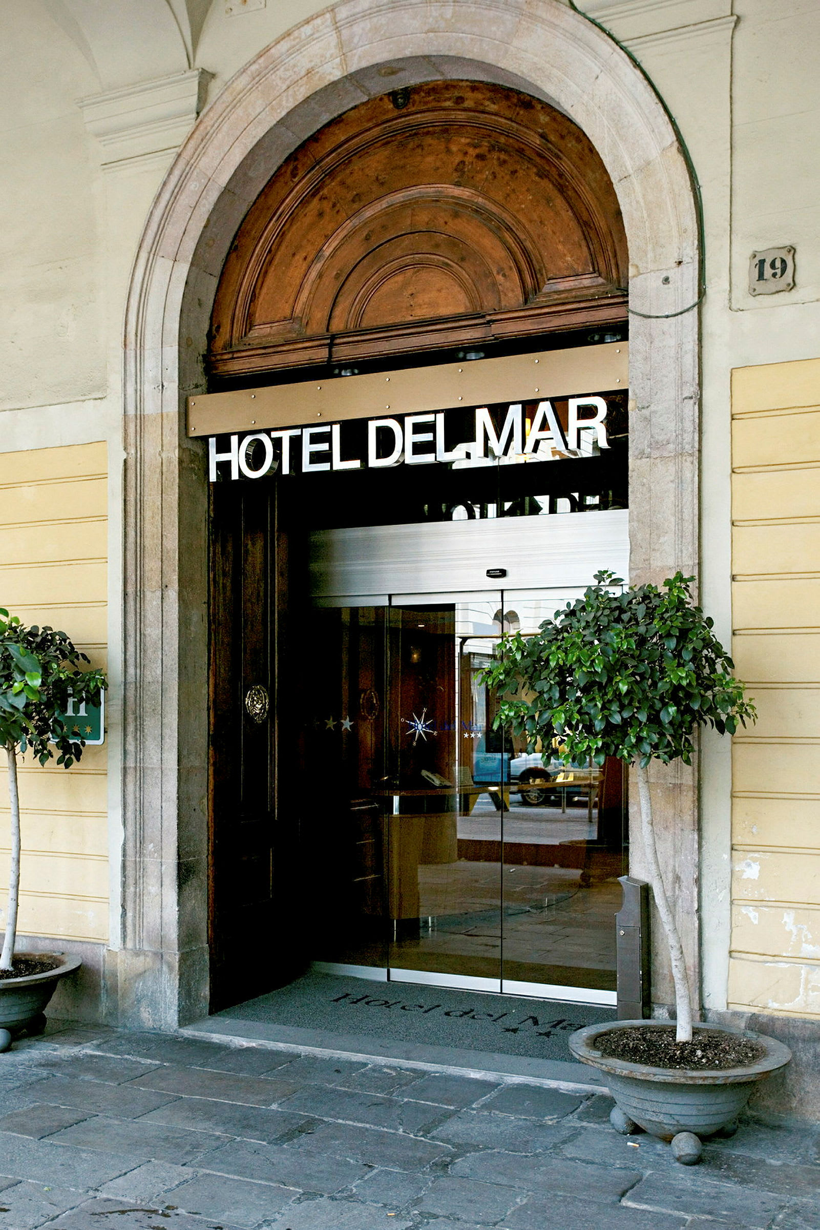 En ingång till ett hotell märkt med texten 'HOTEL DEL MAR'. Entrén är välvd med glasdörrar och har dekorativa blomkrukor på vardera sidan. (AI-genererad text)