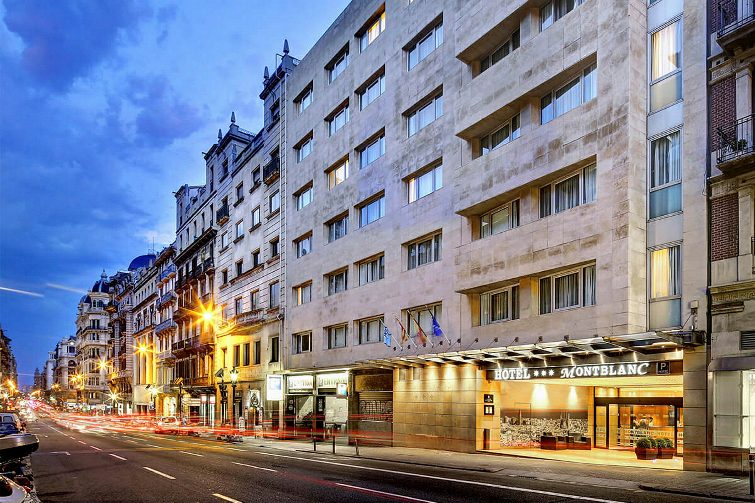 En stadsgata med ett hotell som heter Montblanc syns på högra sidan, med flerfasadiga byggnader och kvällsbelysning. (AI-genererad text)
