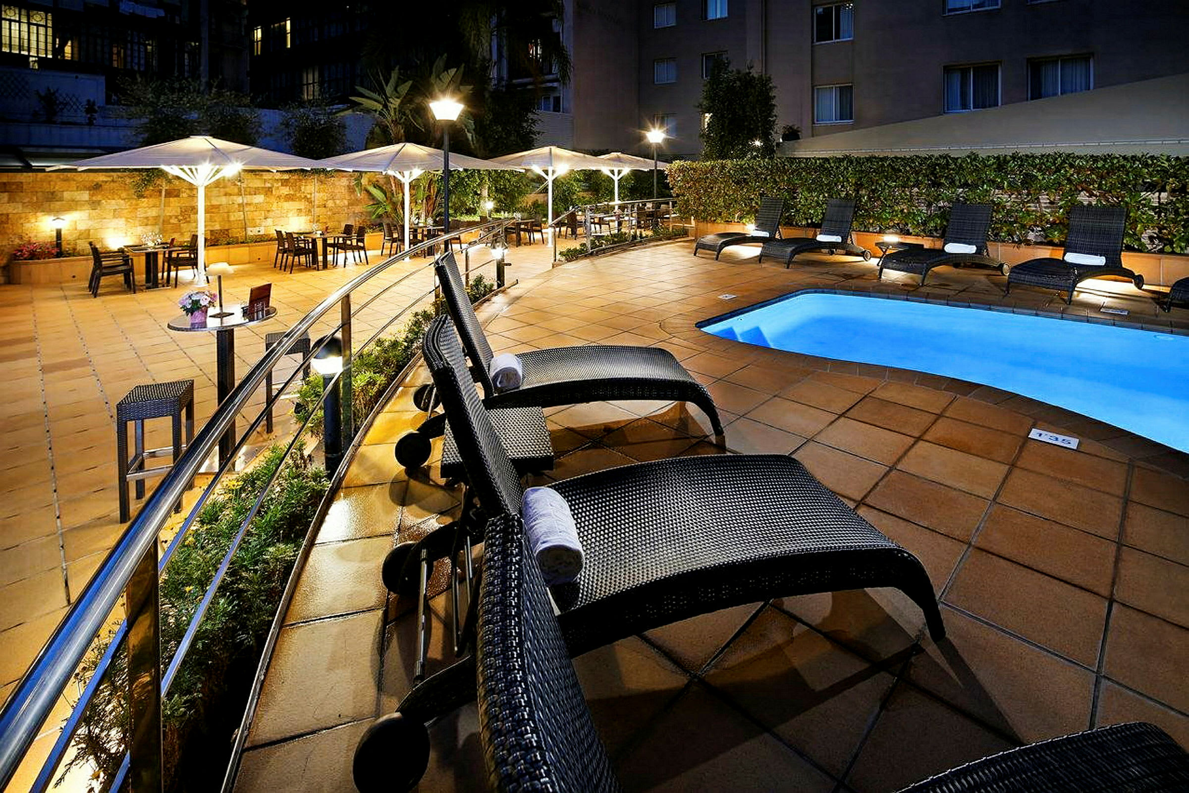 Hotellets poolområde med solstolar och parasoll vid ett upplyst och kaklat uteområde. (AI-genererad text)