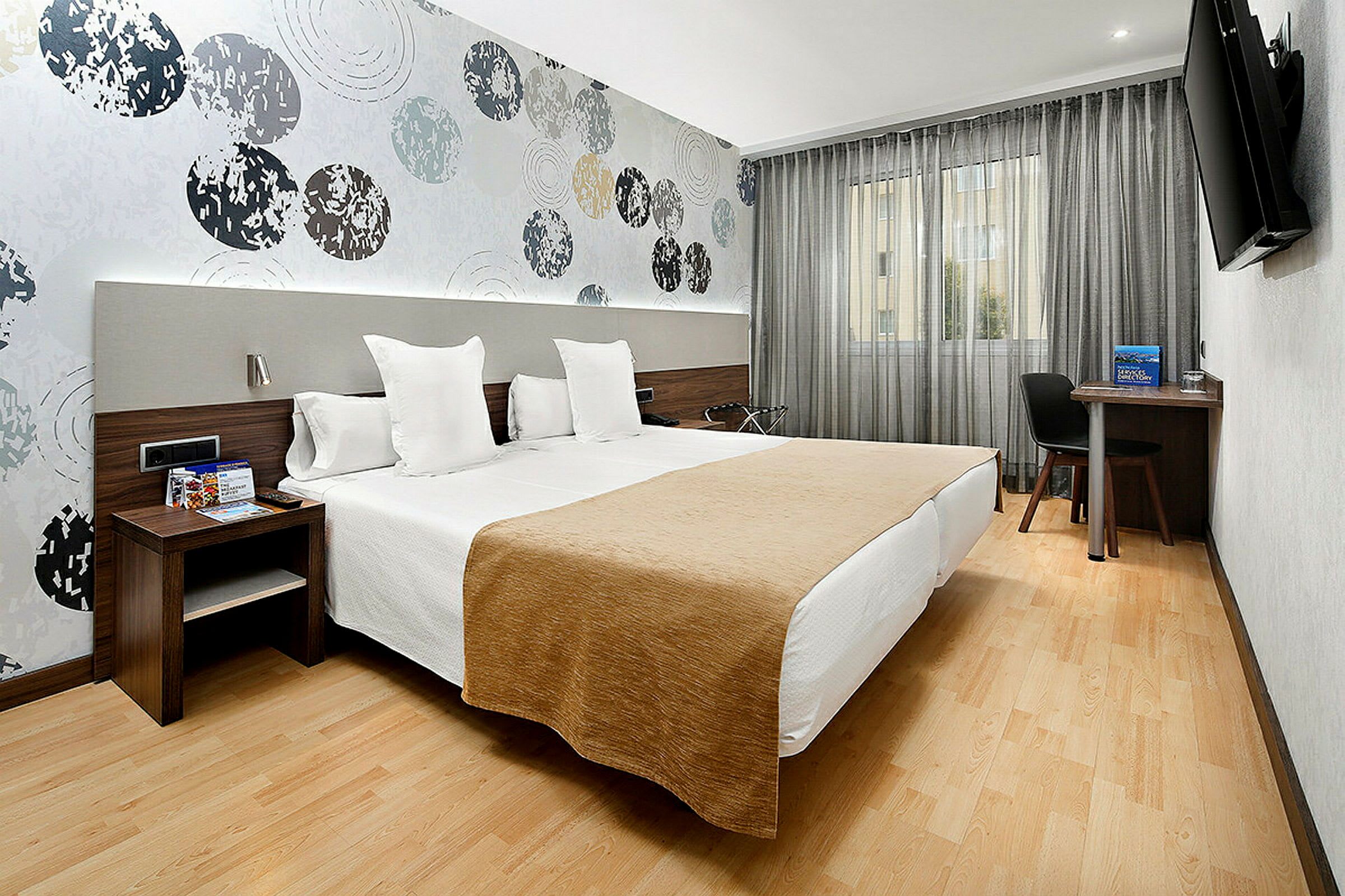 Ett hotellrum med två enkelsängar placerade bredvid varandra. Väggarna är dekorerade med ett mönster av cirklar, och det finns en tv, skrivbord och stol vid fönstret. (AI-genererad text)