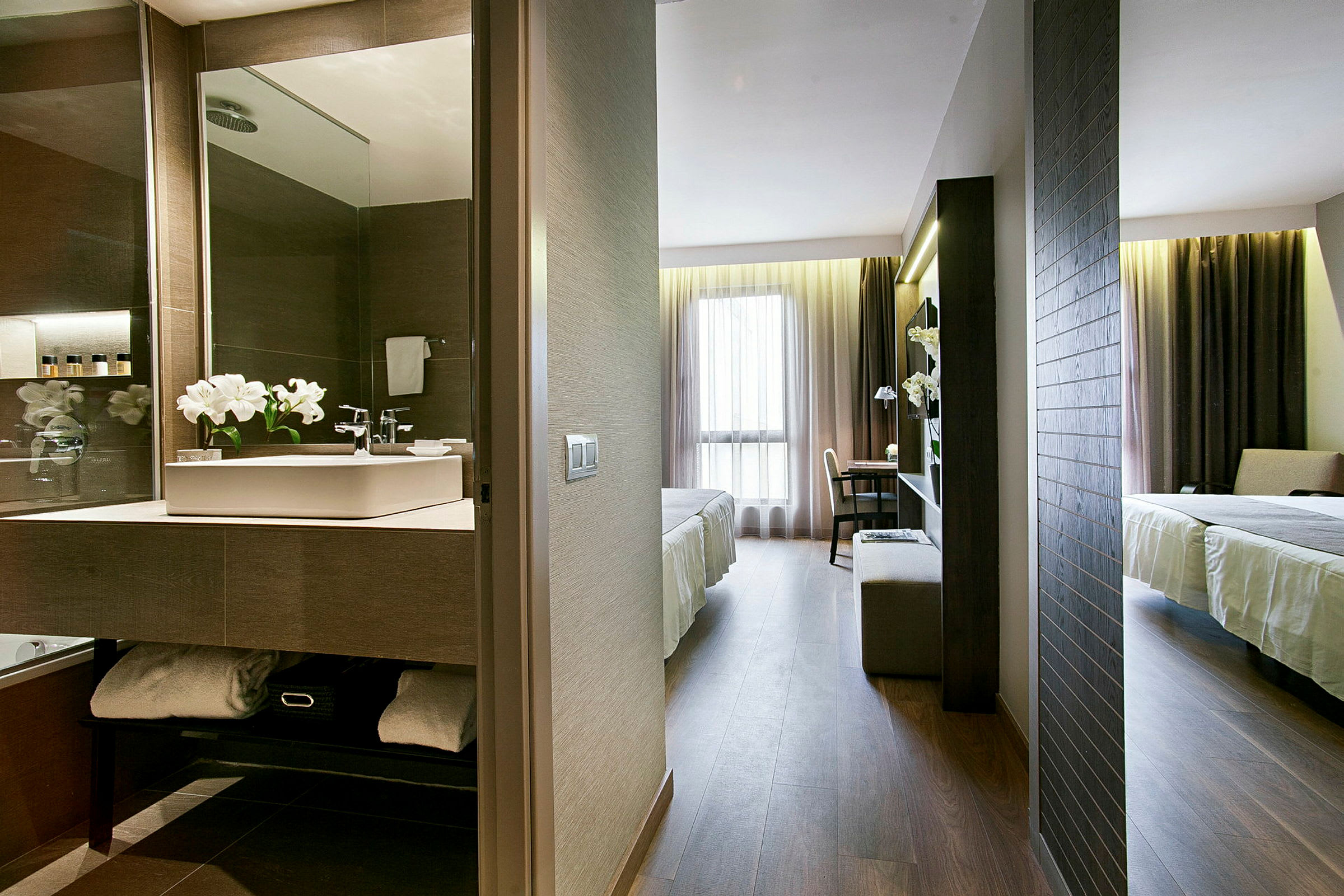 Bild av ett hotellrum med utsikt mot ett badrum och sovrummet. Badrummet har en spegel med ljus, handfat och blommor, medan sovrummet har en bädd och ett skrivbord vid ett stort fönster. (AI-genererad text)