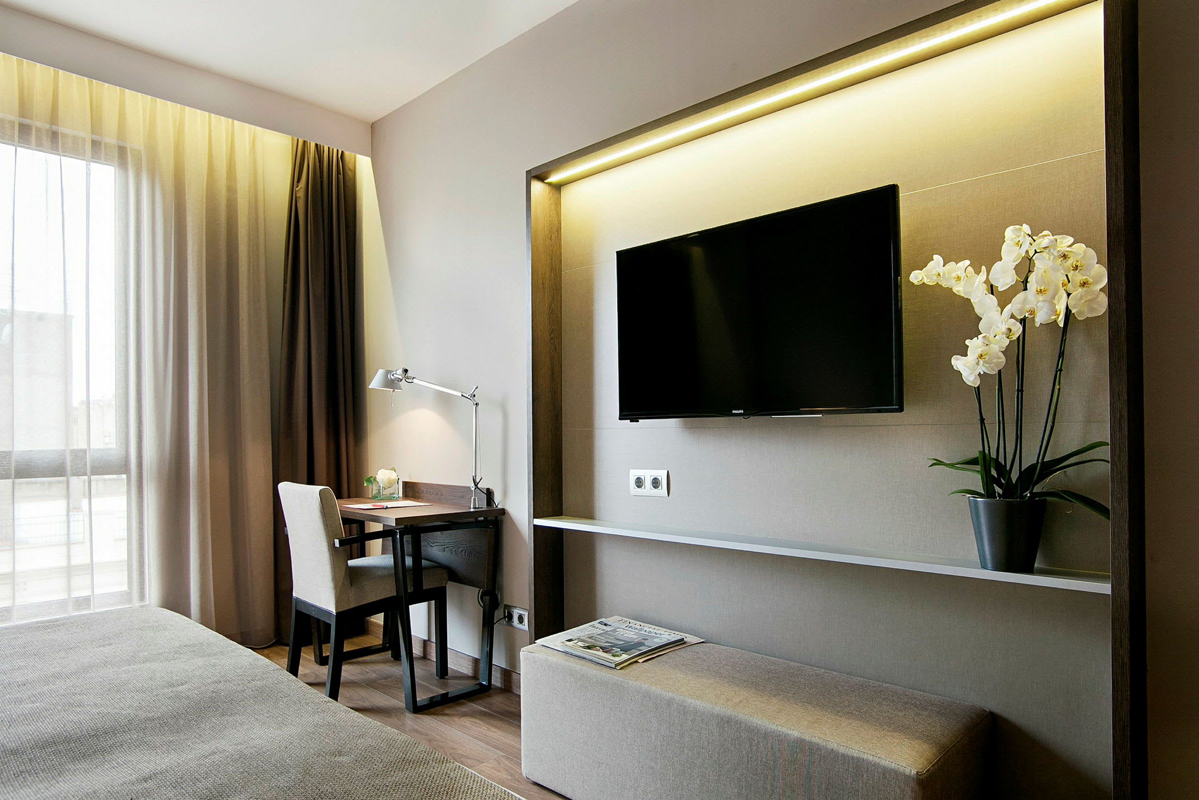 Bild av ett modernt inrett hotellrum med skrivbord, stol, lampa, väggmonterad TV och en blomkruka med vita orkidéer. (AI-genererad text)