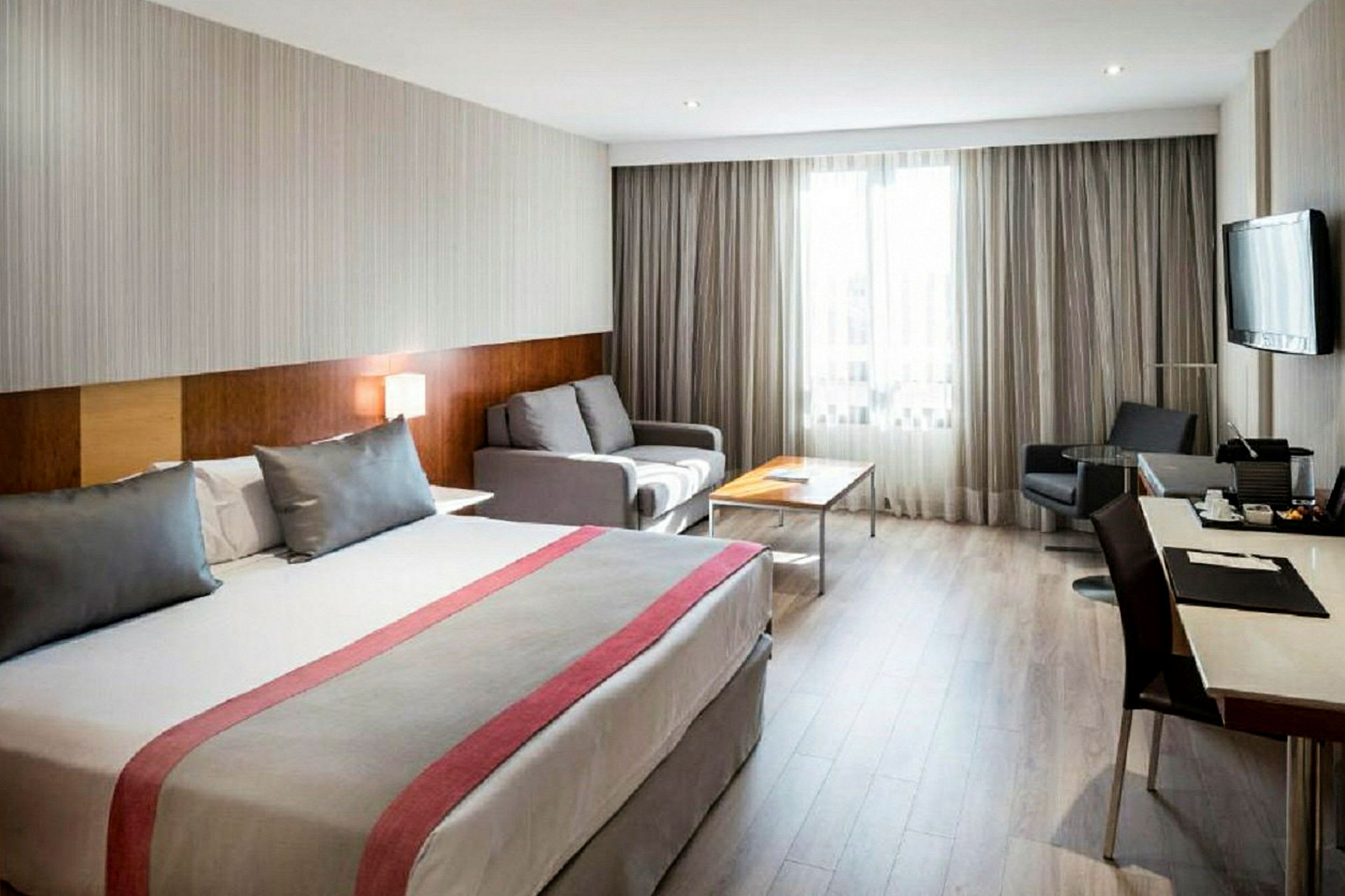 Ett hotellrum med en stor säng, en soffa och ett skrivbord. Ett fönster med gardiner släpper in ljus. (AI-genererad text)