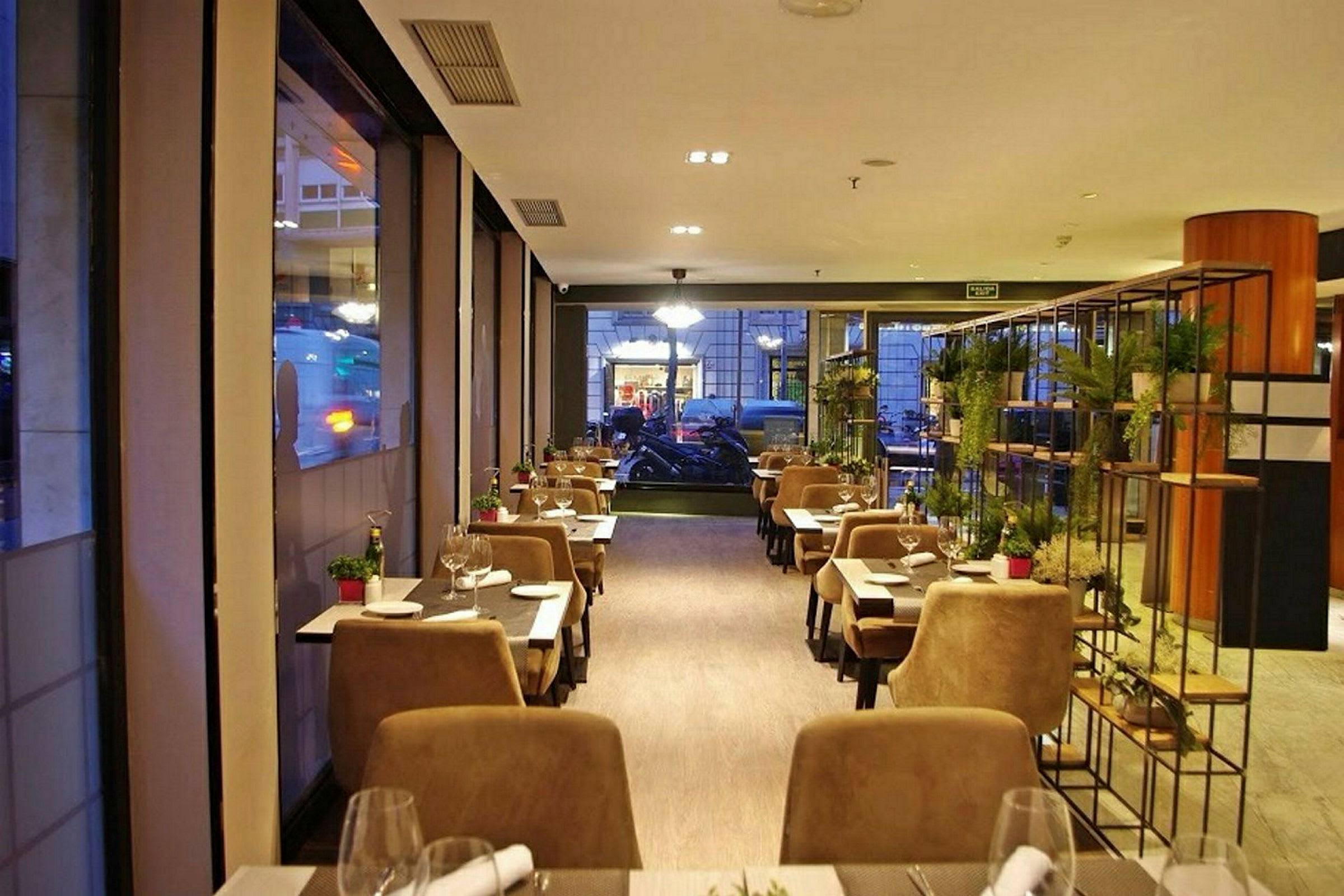 En mysig restaurang med bord och stolar arrangerade längs fönster och ett dekorativt ställ med växter. (AI-genererad text)