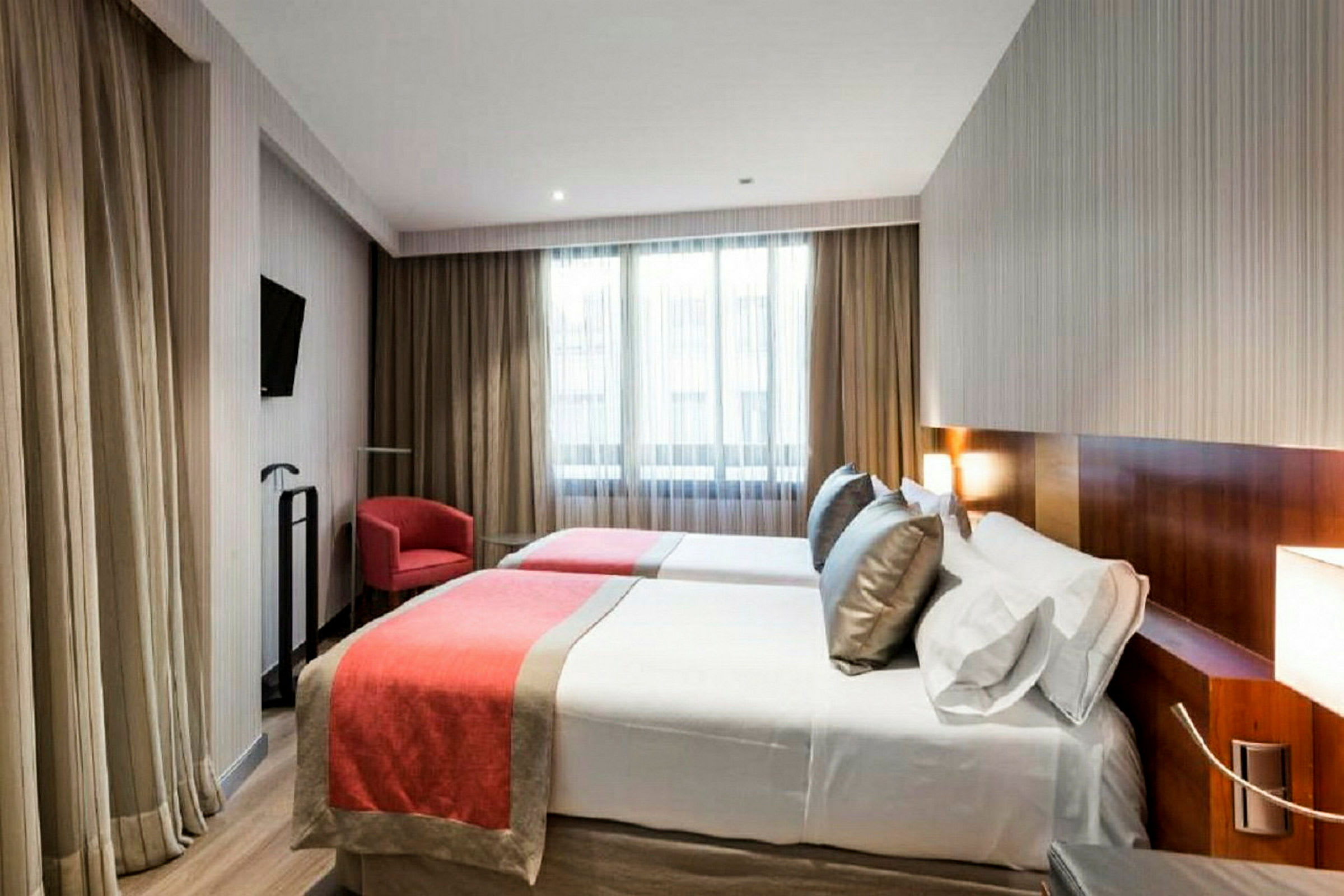 Ett hotellrum med två enkelsängar bäddade med röd-orange överkast och vita lakan. En röd fåtölj och ett litet glasbord syns bredvid ett fönster med tunna gardiner. (AI-genererad text)