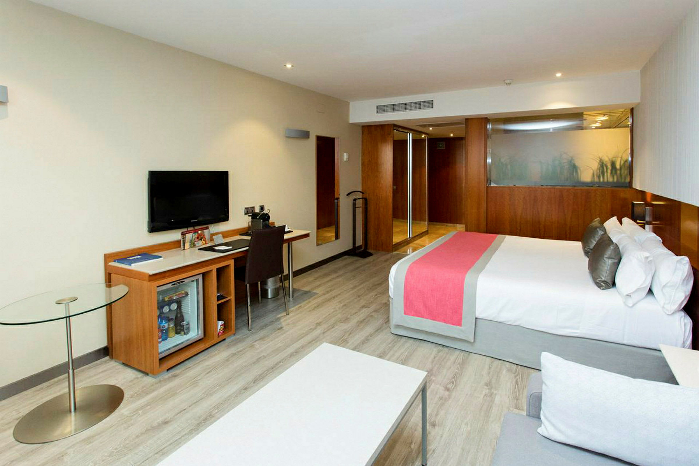 Ett modernt hotellrum med en dubbelsäng, arbetsbord, TV, och glasbord. (AI-genererad text)