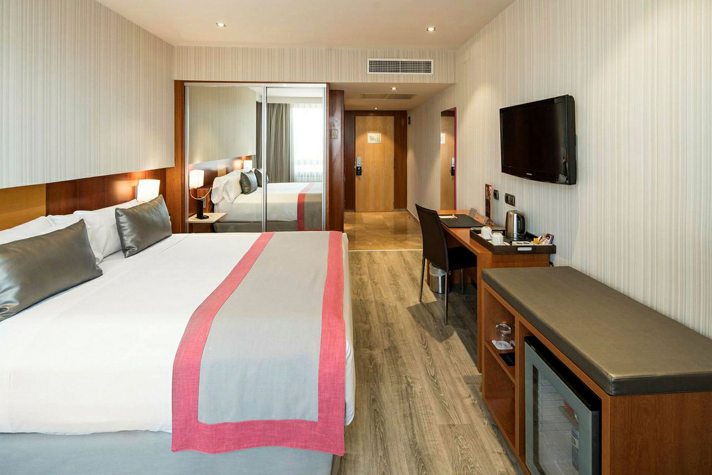 Hotellrum med dubbelsäng, skrivbord, TV på väggen och en garderob med speglar. (AI-genererad text)