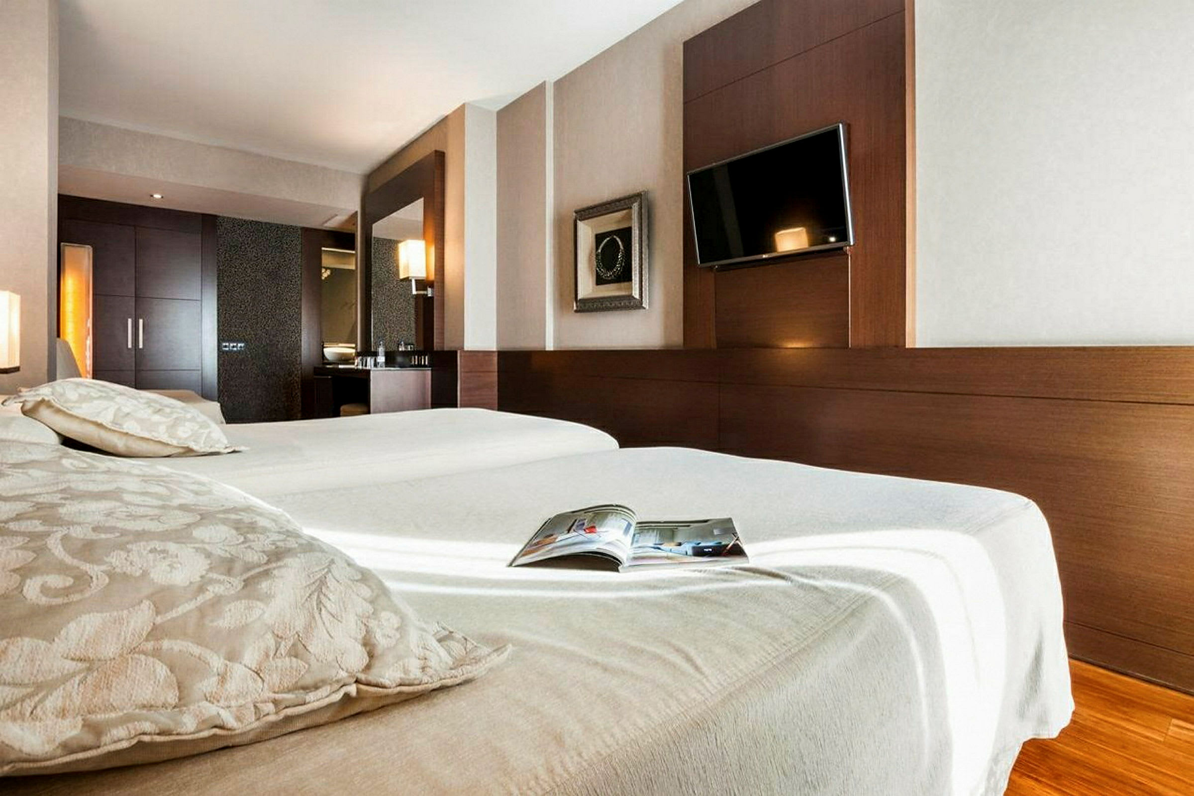 Ett ljust hotellrum med en stor säng, en väggmonterad TV och en spegel. Ett öppet magasin ligger på sängen. (AI-genererad text)