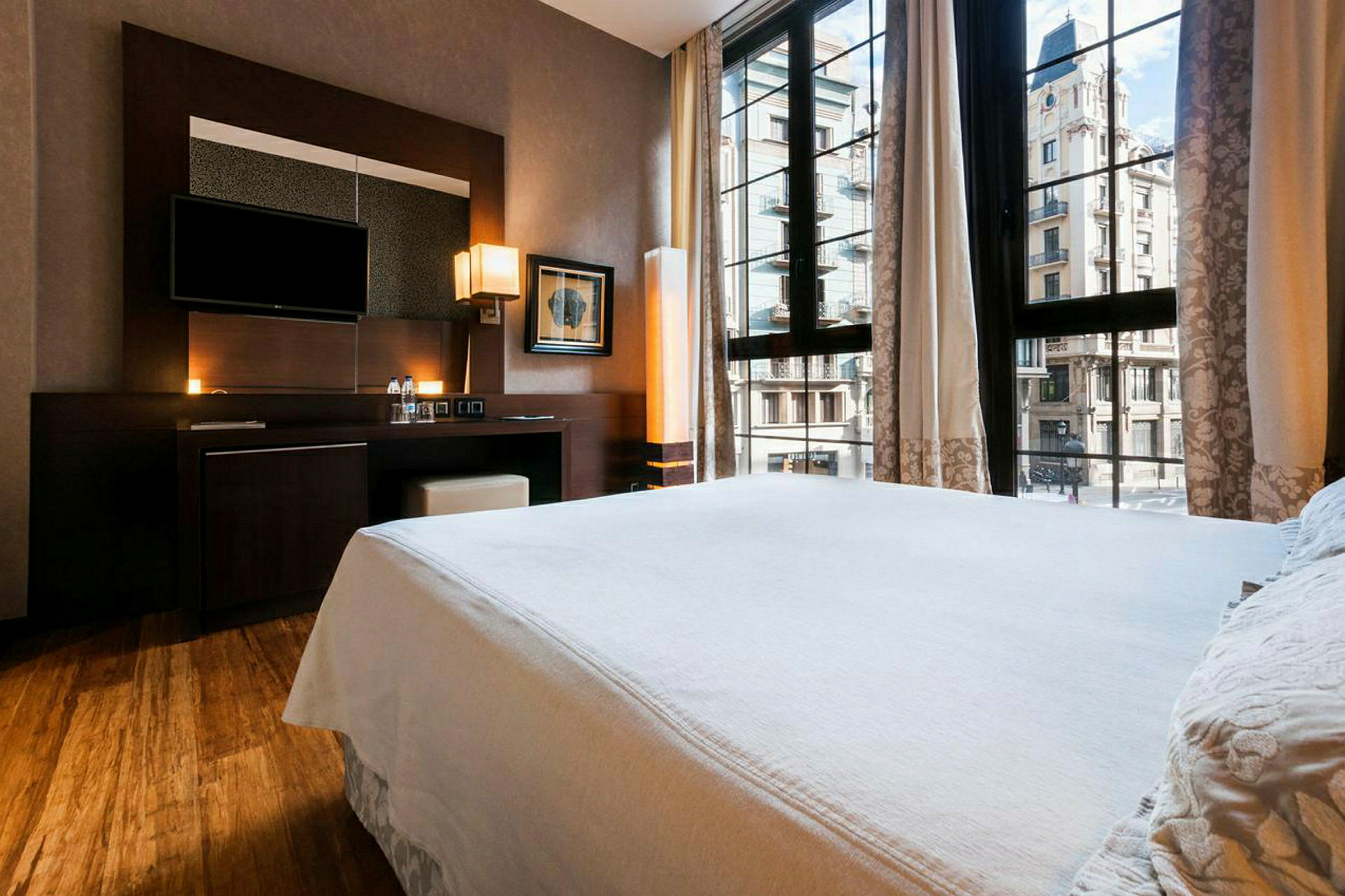 Ett hotellrum med en bred vit säng framför ett stort fönster med utsikt mot en stadsgata och byggnader. (AI-genererad text)